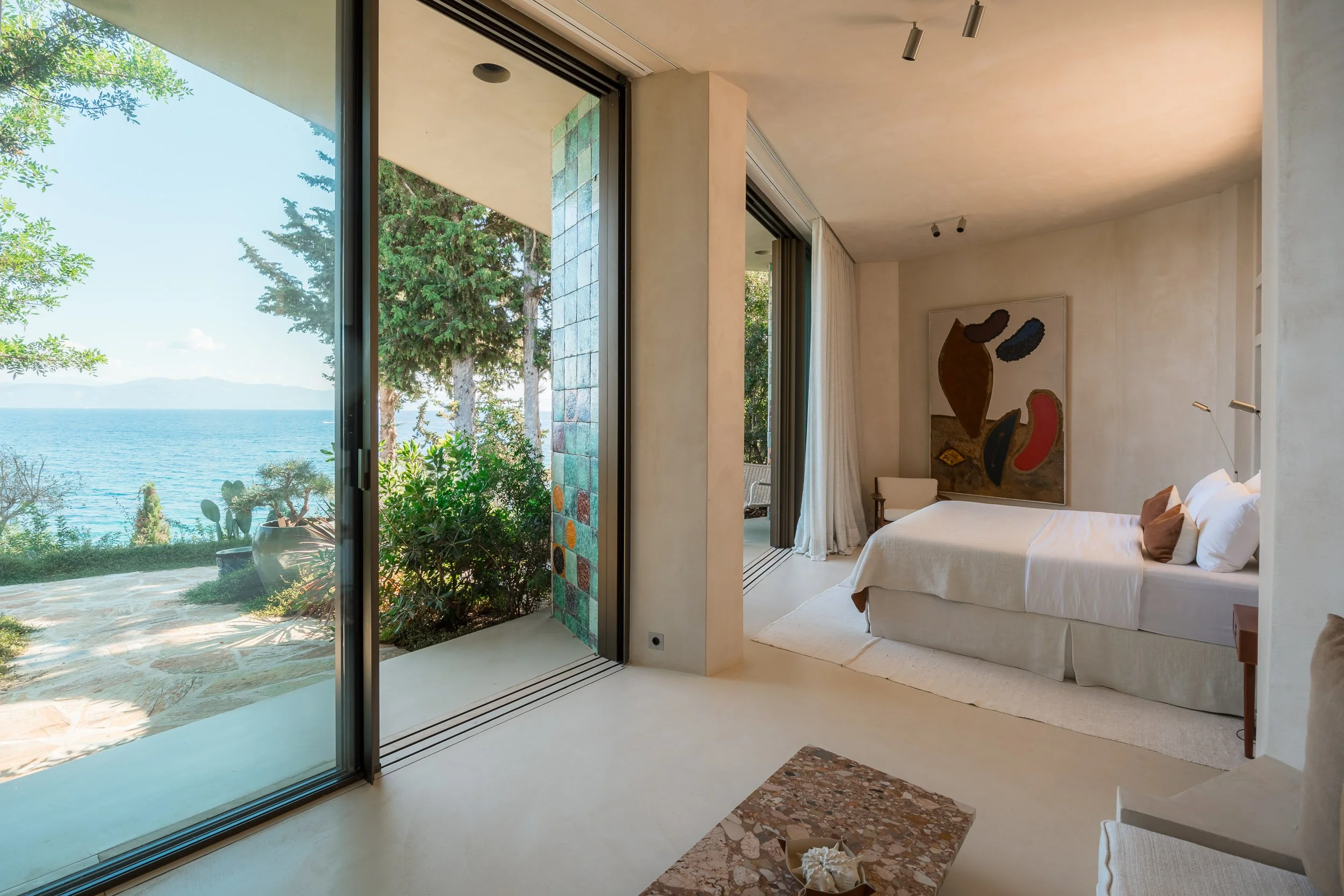 A bedroom with a large glass sliding door leading to a balcony with a view of trees, bushes, and the ocean in the background. The room has minimal decor, a white bed with brown and white pillows, a piece of abstract art on the wall, and a small table
