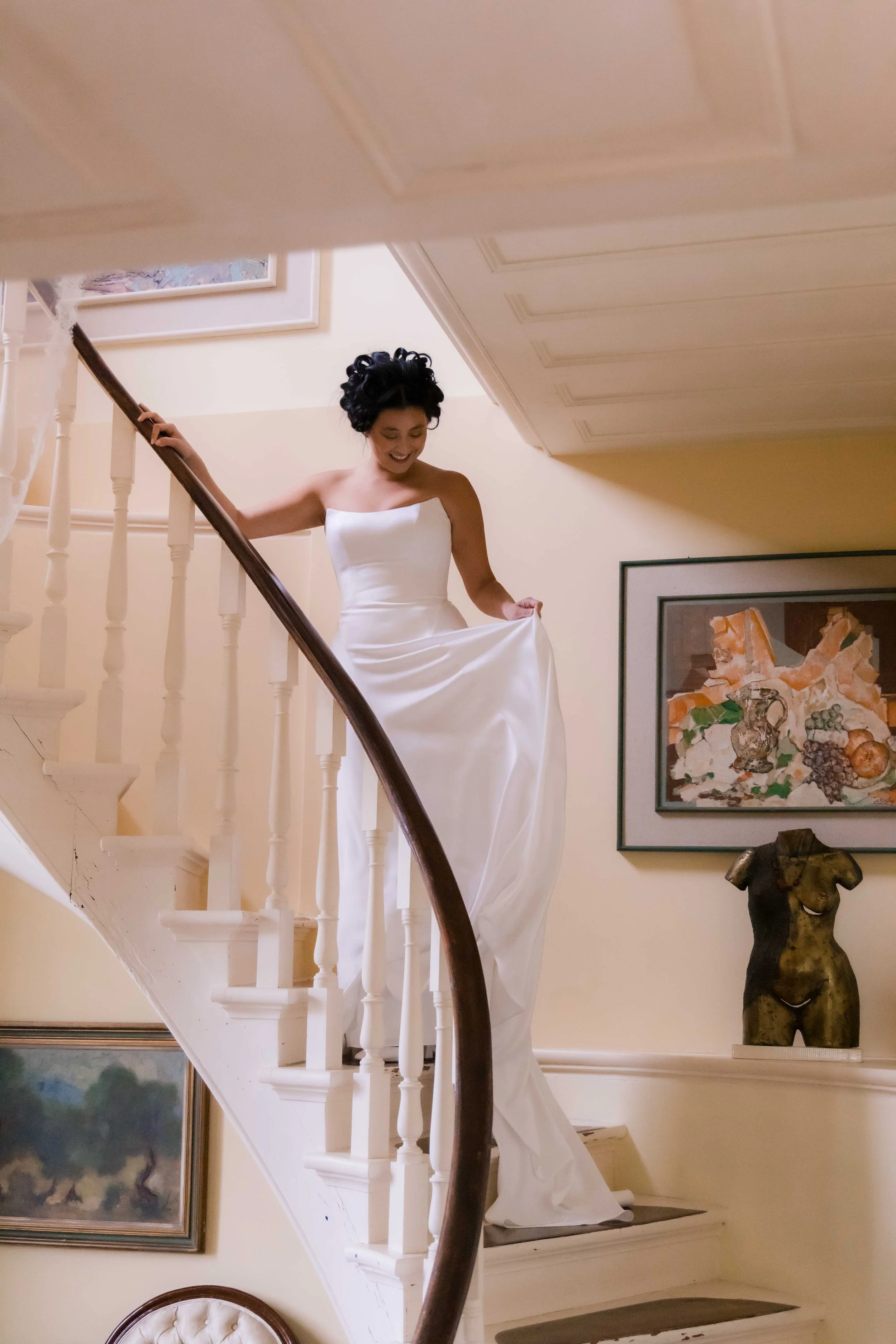 A woman in a white wedding dress standing on a staircase, holding up part of her dress and smiling, with framed artworks and sculptures on the wall behind her.