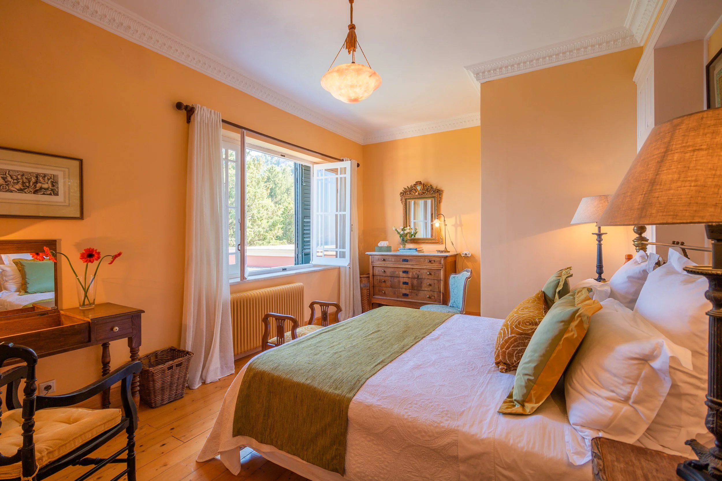 A cozy bedroom with a large bed, pillows, and a green bed runner. There are wooden furniture pieces including a dressing table with a mirror, a dresser, and chairs. The room is decorated with lamps, wall art, and flowers. An open window lets in natur