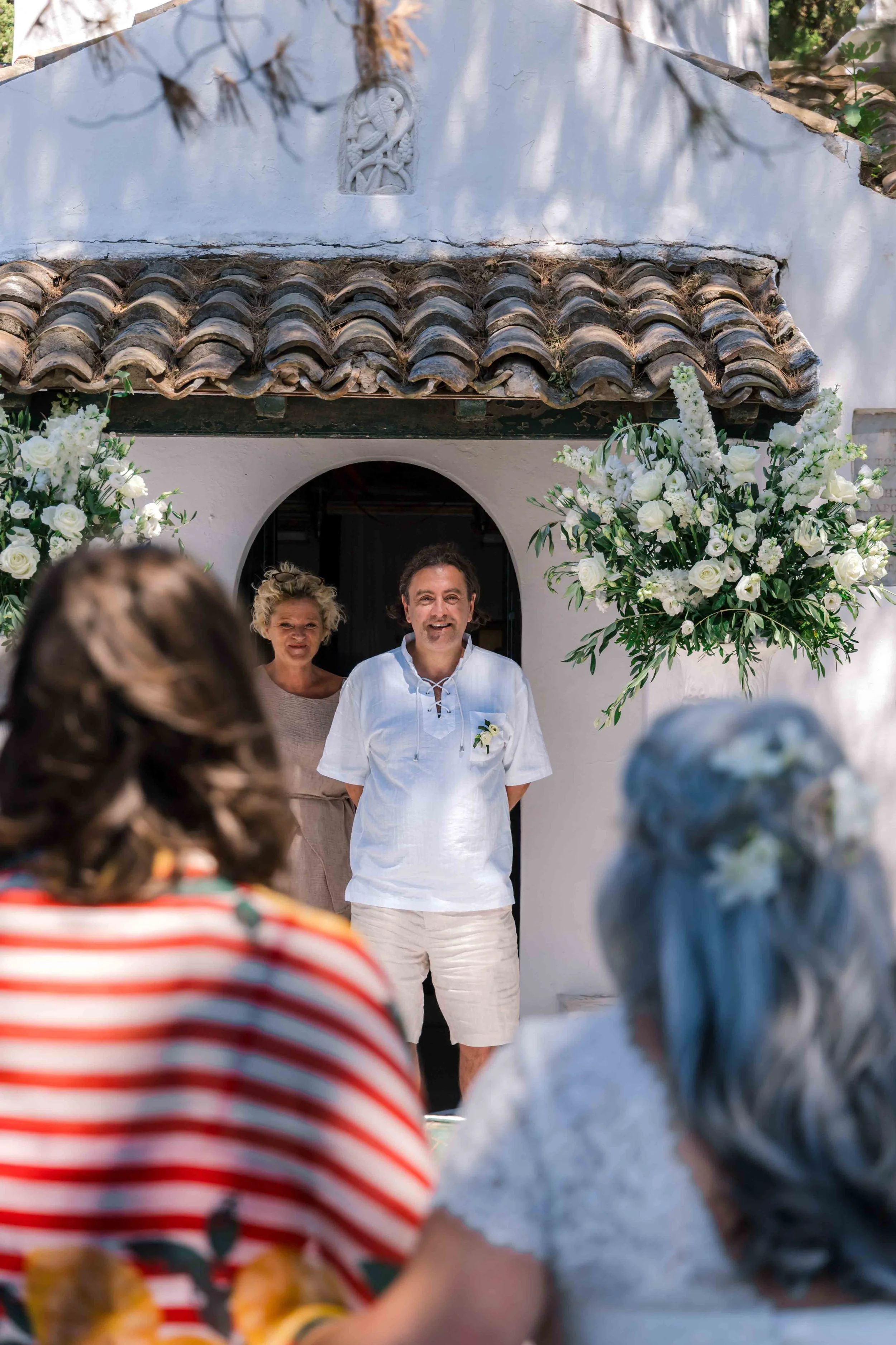 A couple stands in front of an arched doorway decorated with large white flower arrangements, during an outdoor wedding ceremony, with guests in the foreground.