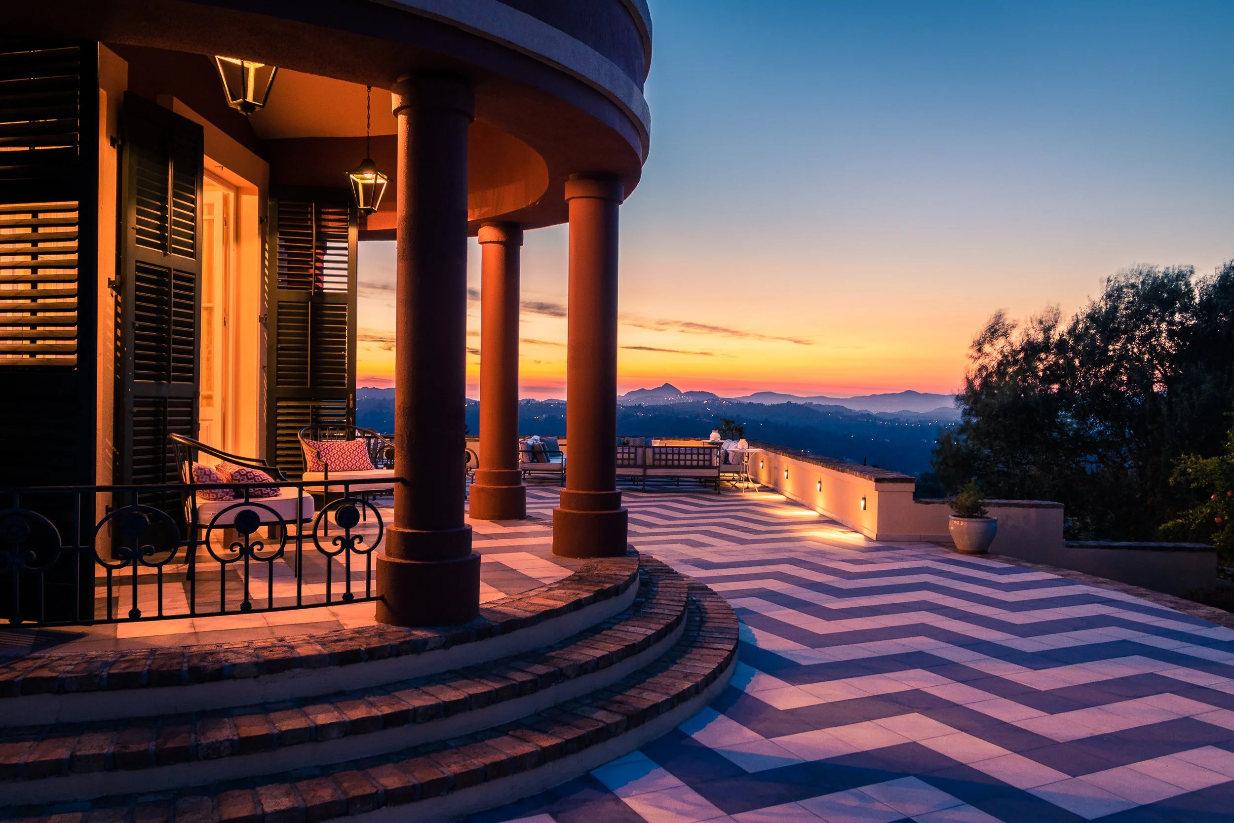 Luxury house terrace with patterned tile floor, large columns, and outdoor seating, overlooking a scenic mountain sunset with colorful sky.