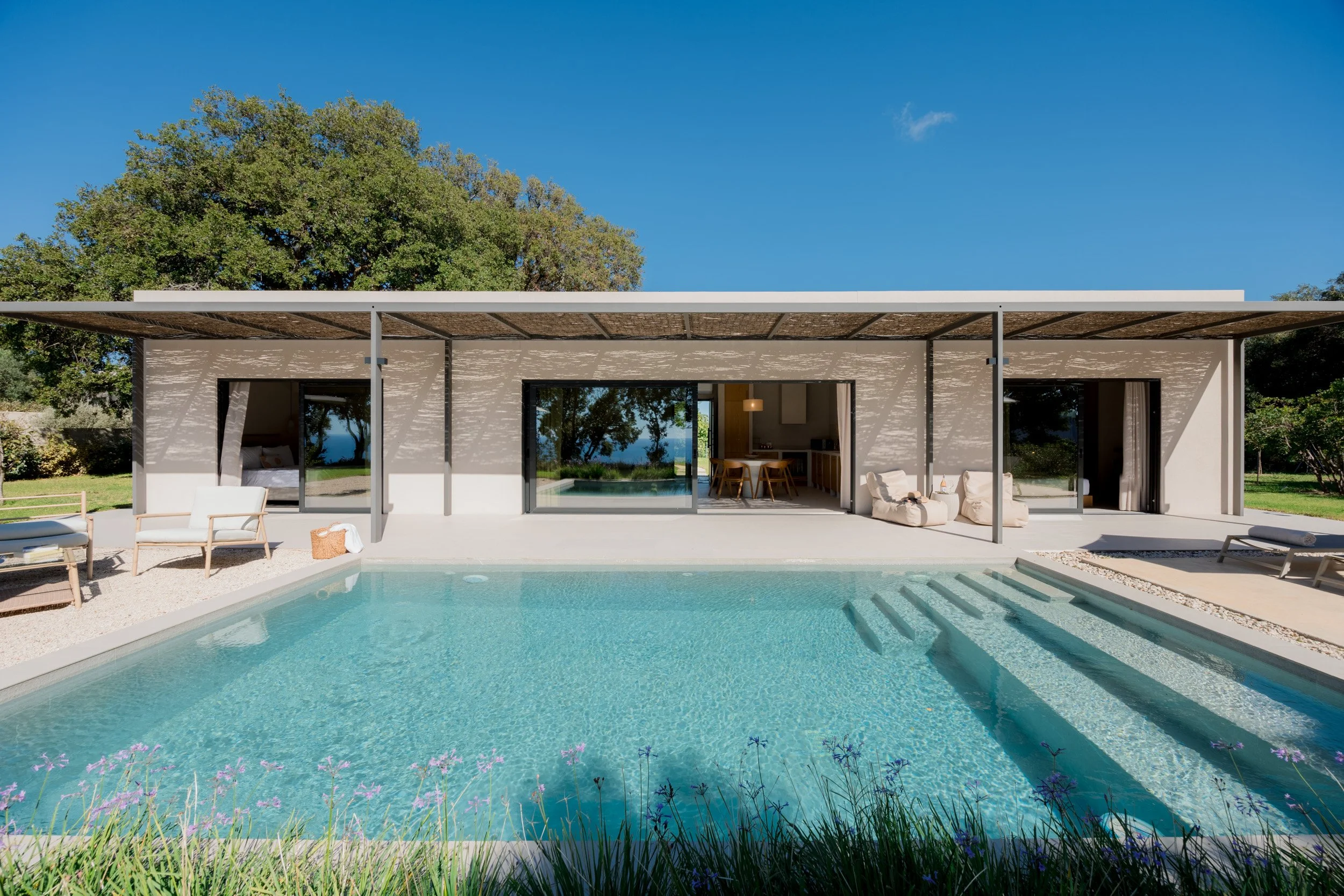 Modern house with large sliding glass doors opening to a swimming pool, outdoor chairs, and a shaded patio with trees in the background.