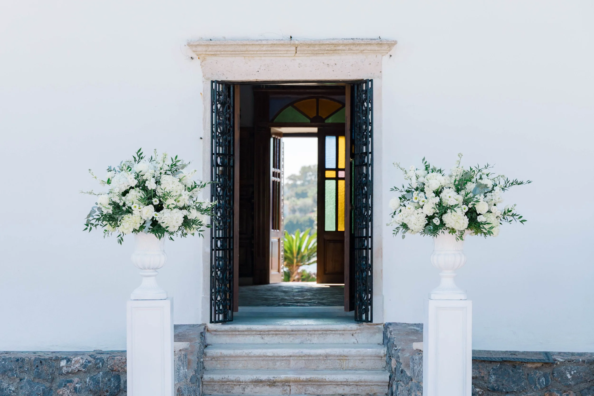 Open door of a white building with two large white floral arrangements on each side, leading to a view outside with greenery.