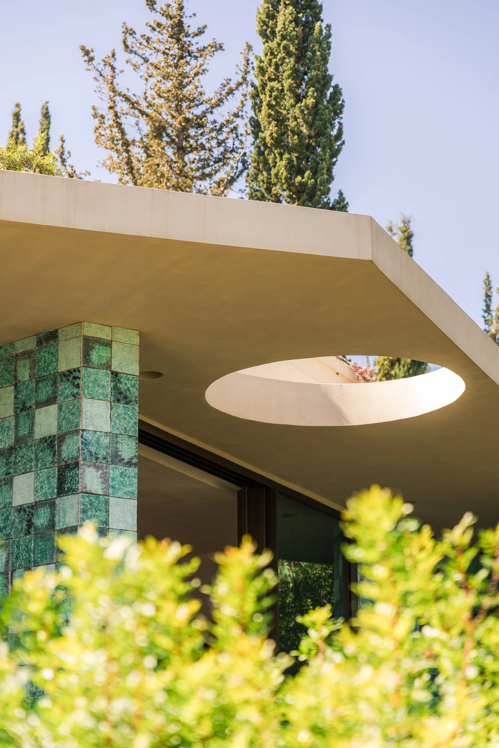 Close-up of a modern building's ceiling with a circular skylight, green-tiled wall, large glass window, and surrounding greenery including trees and bushes.
