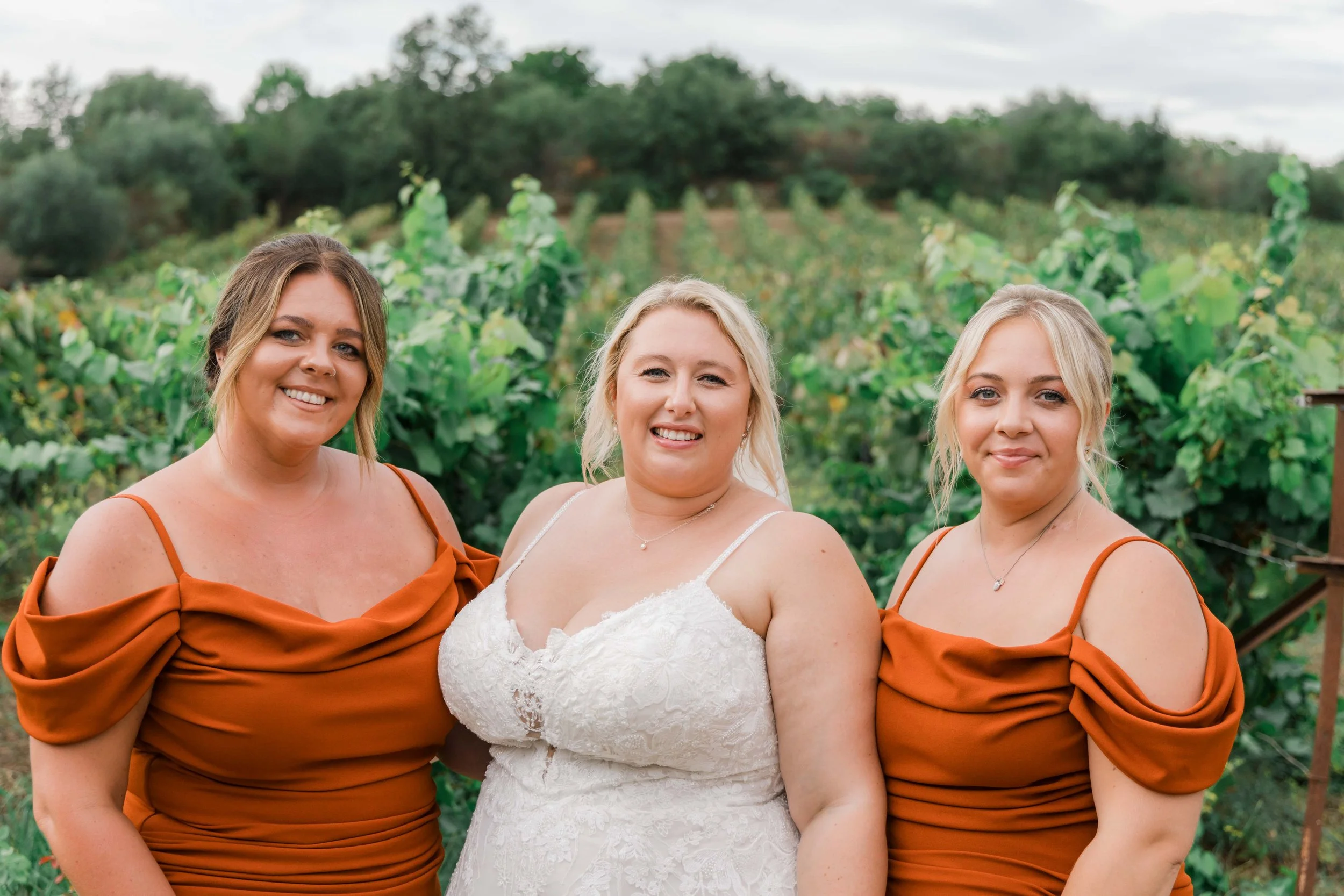 A bride in a white lace wedding dress and two bridesmaids in matching rust-colored dresses standing outdoors in a vineyard.