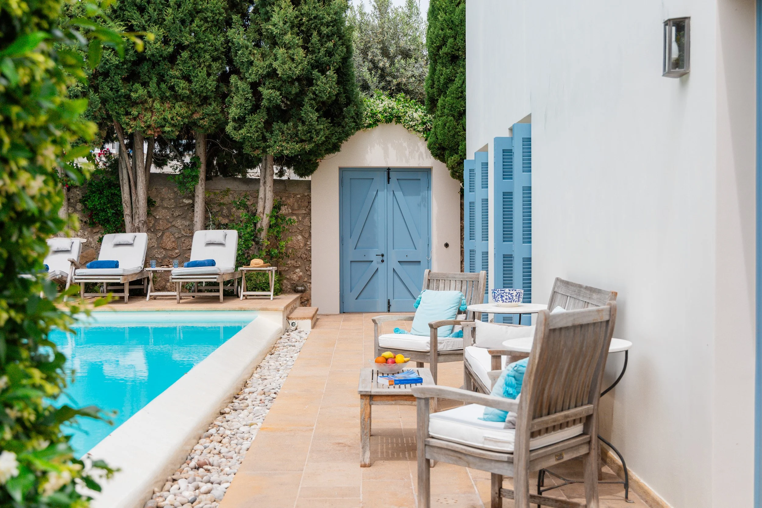 Outdoor poolside area with wooden lounge chairs, blue cushions, small tables, and a white wall with blue shutters, surrounded by green trees and shrubs.