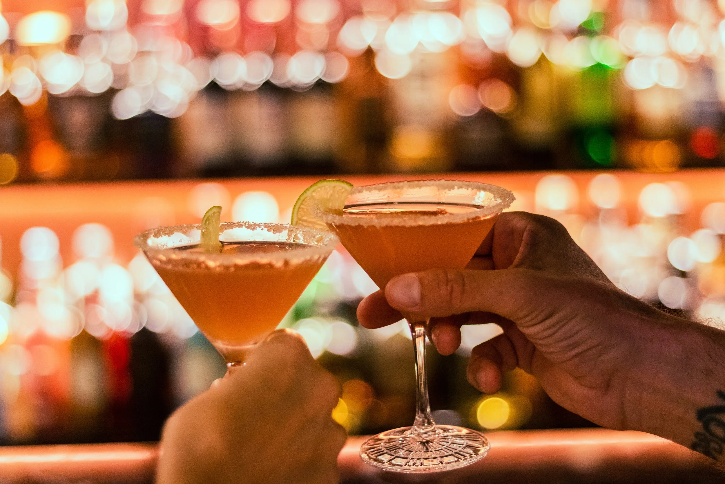 Two hands holding cocktails with salted rims and lime wedges at a bar with colorful blurred lights in the background.