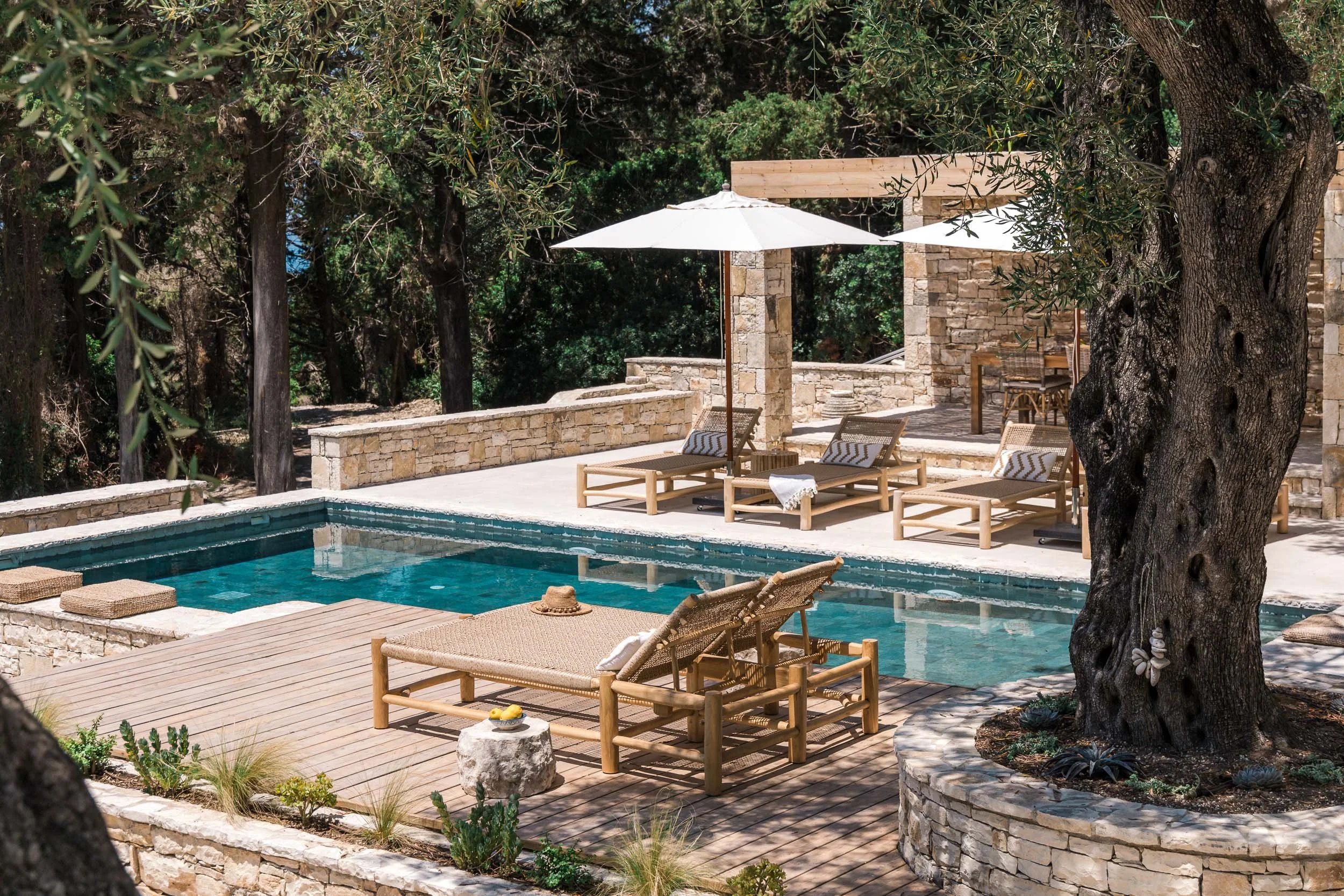 An outdoor pool area with lounge chairs, umbrellas, and a stone patio surrounded by trees.