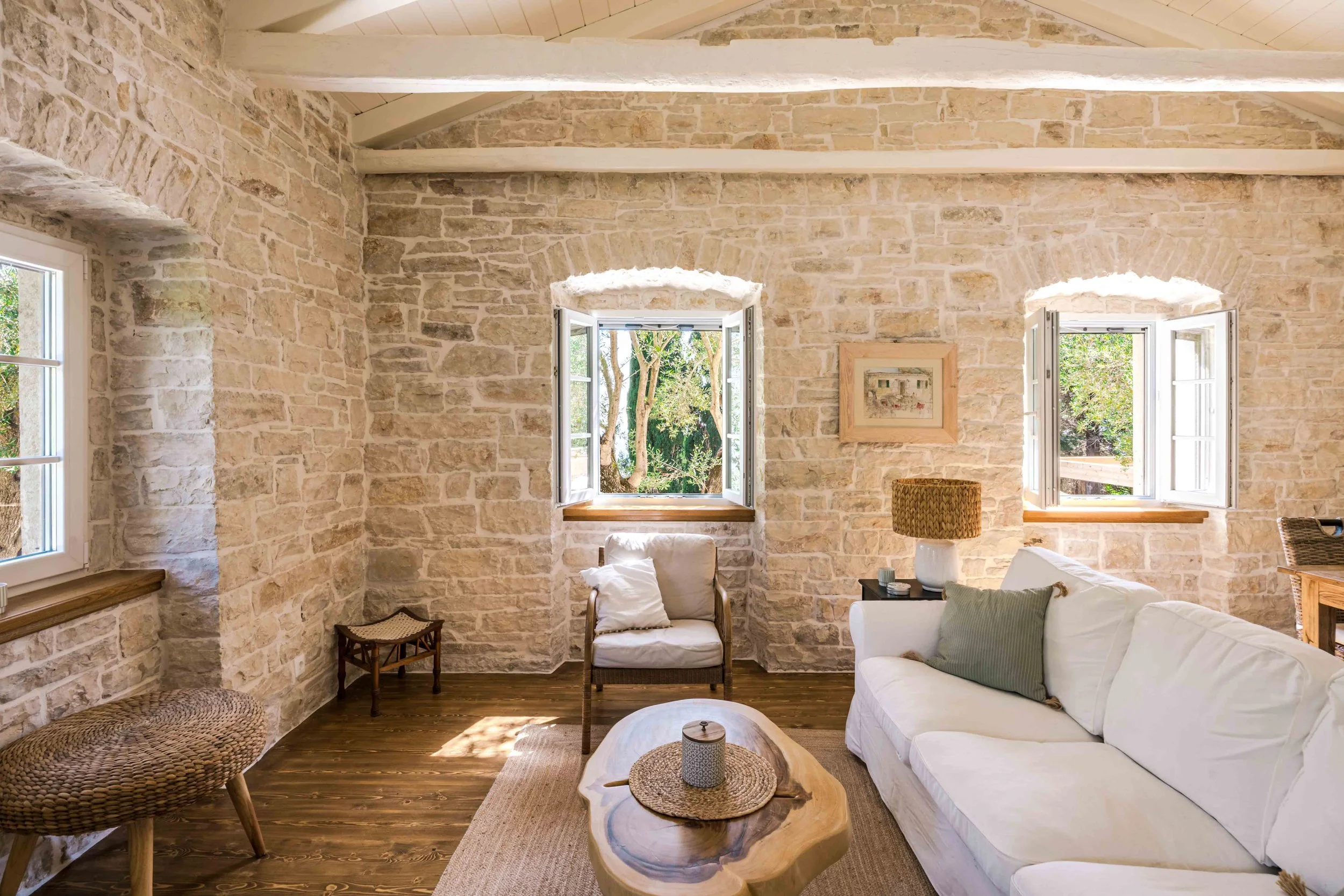 Living room with exposed stone walls, three windows open to outdoor greenery, white sofa, wooden coffee table, cushioned armchair, wicker stools, and a table lamp.