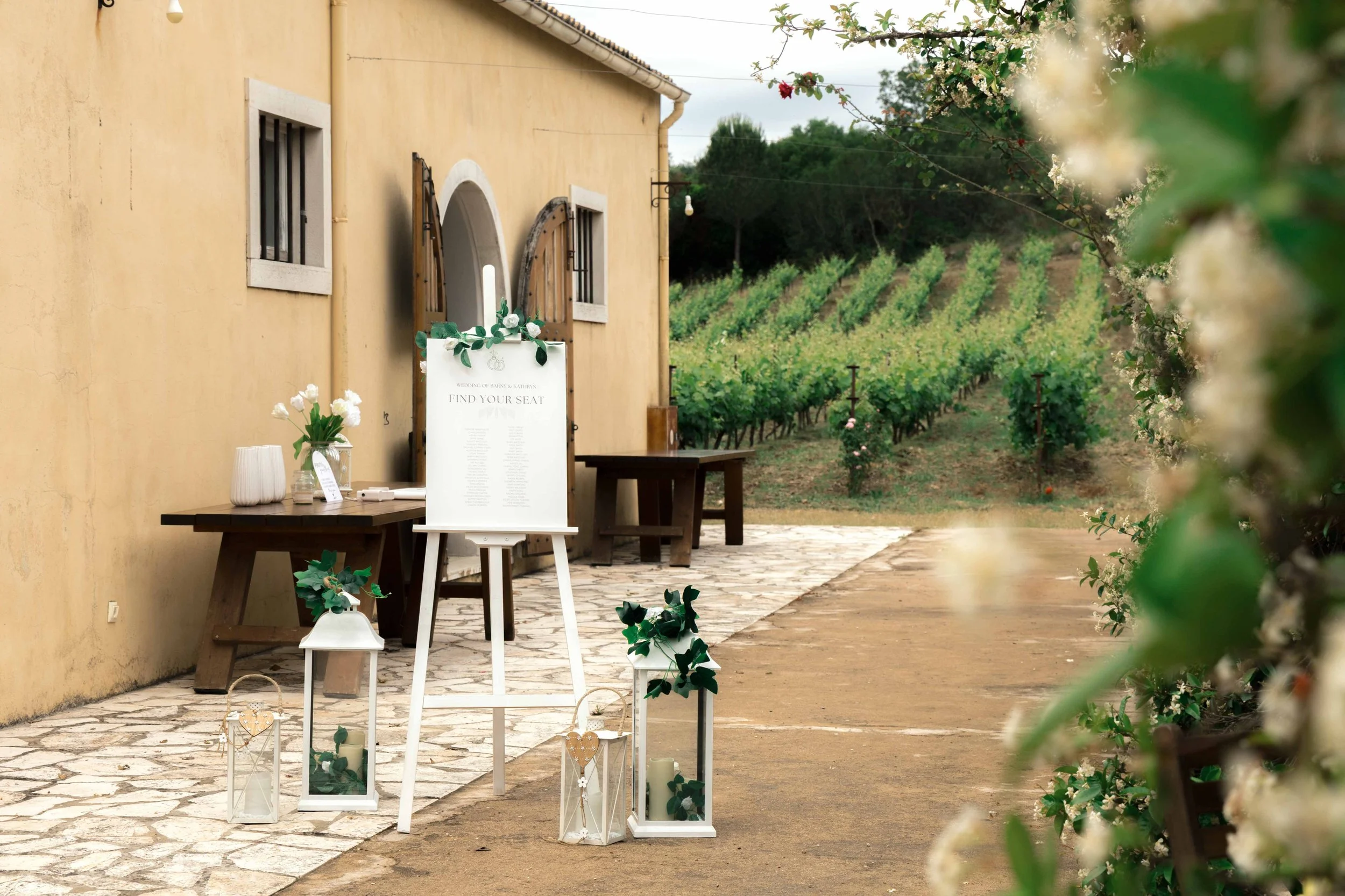 Wedding venue decoration outside with lanterns, flowers, and a sign that says 'Find Your Seat' near a village-style building and a vineyard in the background.