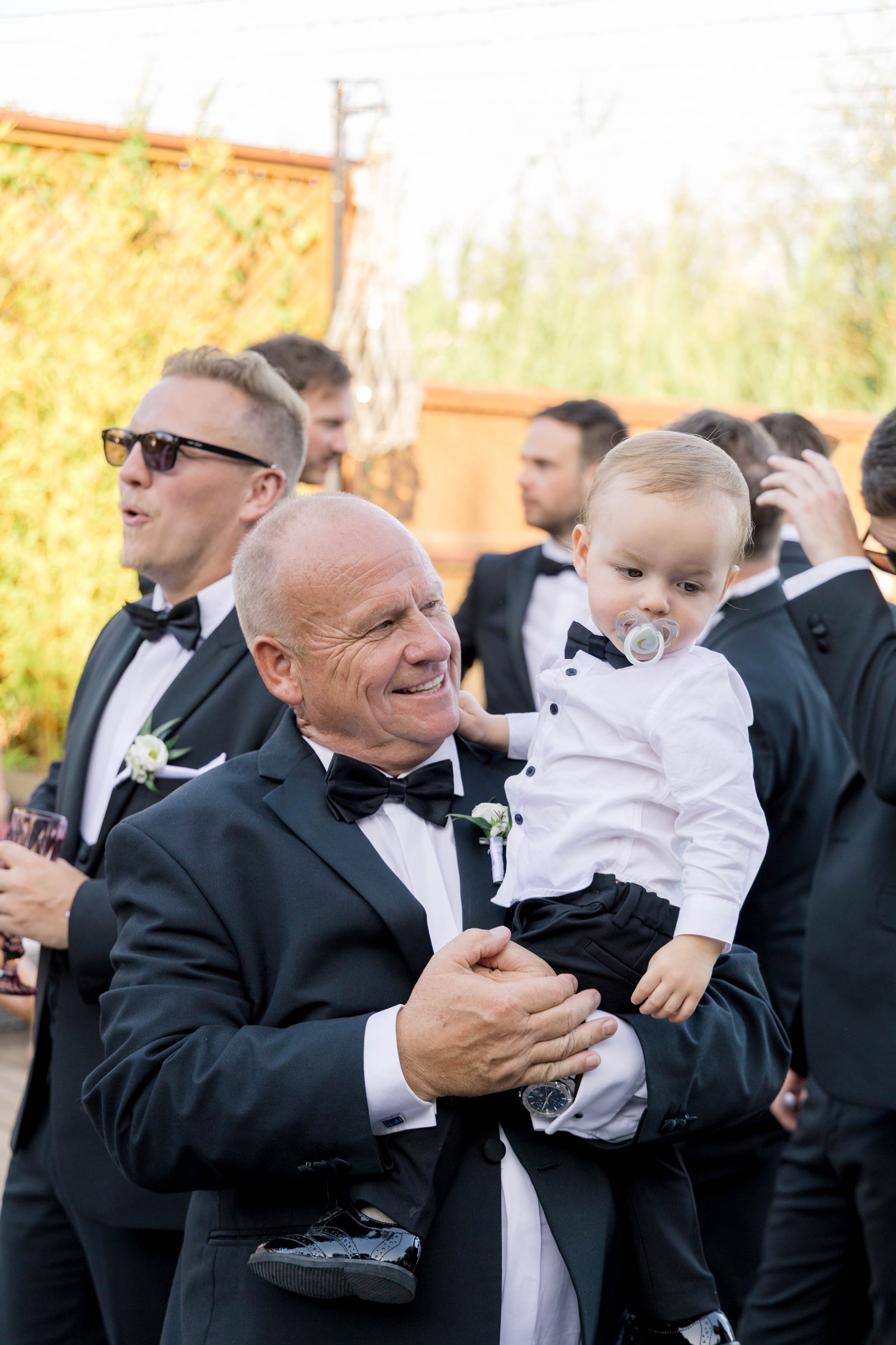 Older man holding a young boy during a formal event, with men in tuxedos in the background.