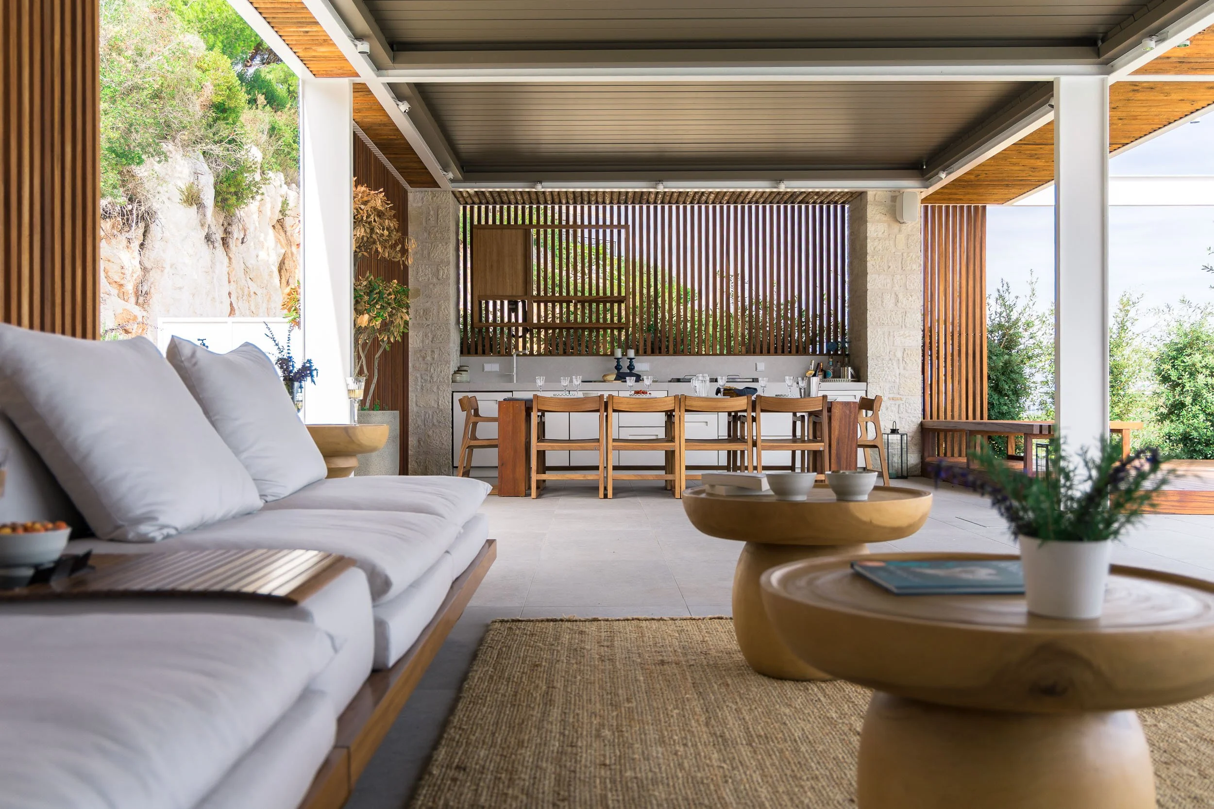 Living area with white sofa, round wooden tables, and a dining table with chairs in the background, all within an open-air space with wooden accents and views of greenery.