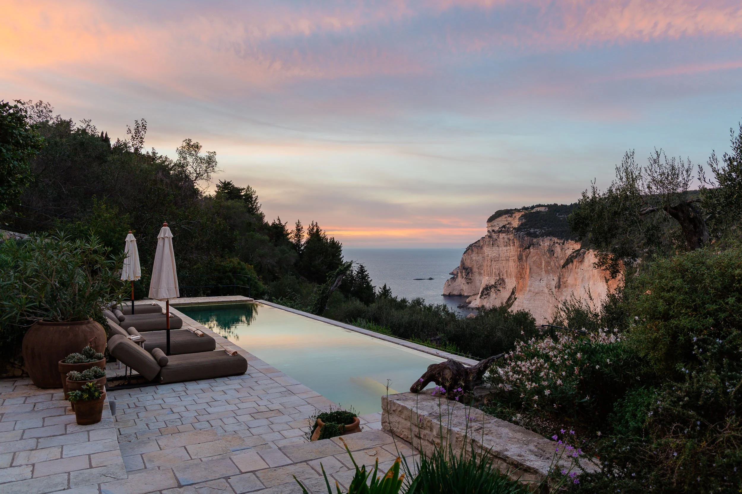 A scenic view of a backyard with a swimming pool, lounge chairs, and umbrellas overlooking a rocky cliff and the ocean at sunset.