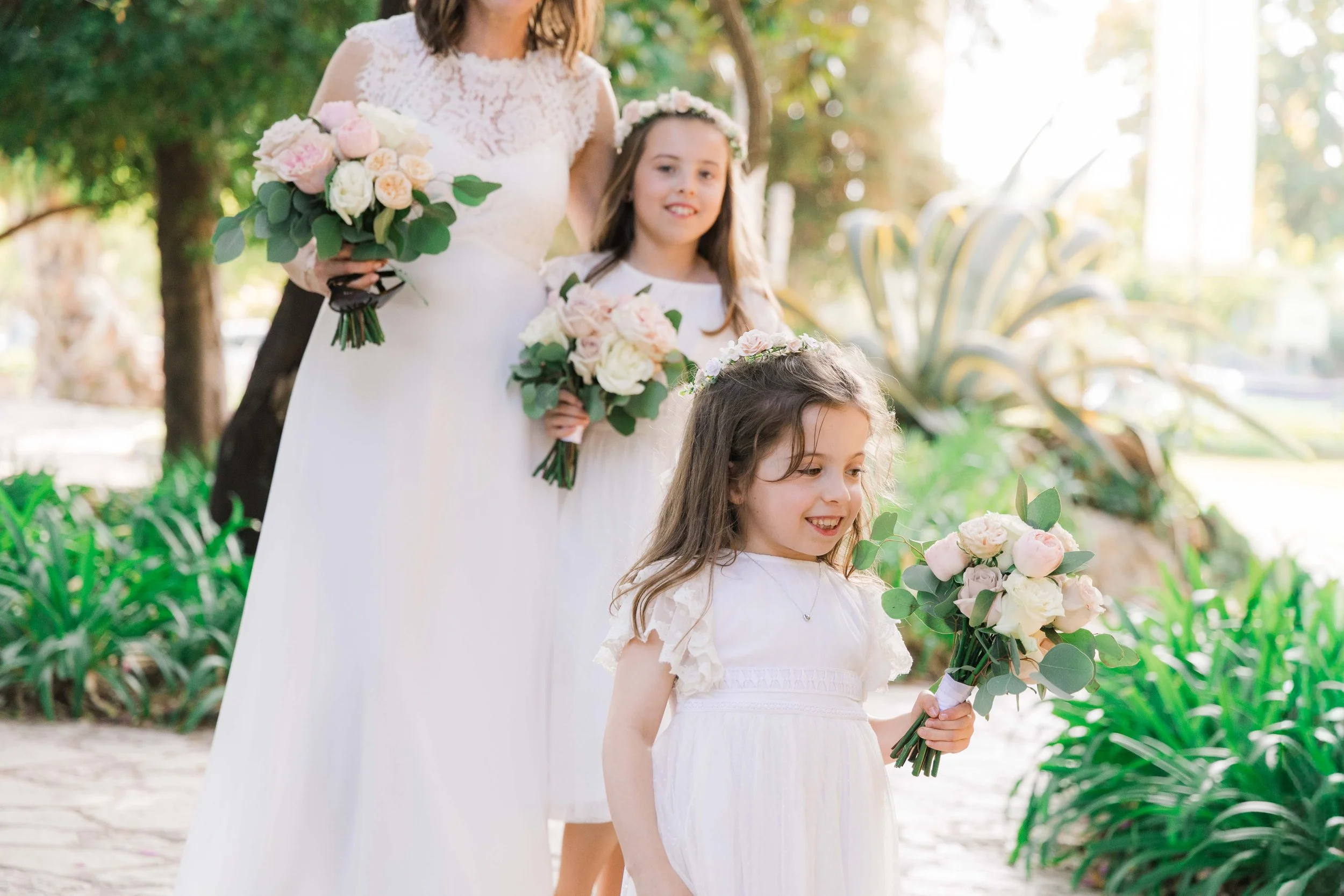 A bride and three young girls, all wearing white dresses and floral crowns, holding bouquets of pink and white flowers outdoors in a garden setting.