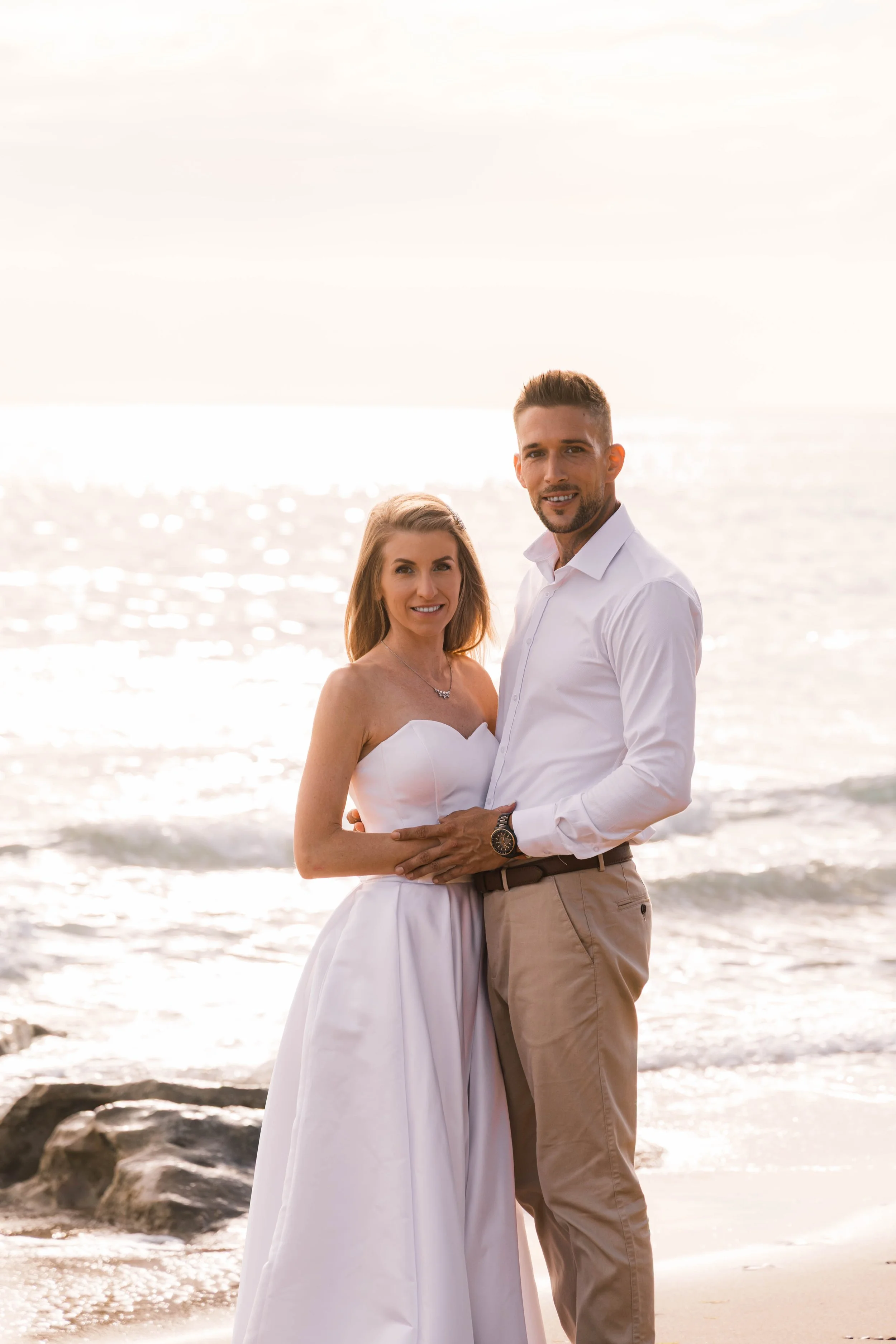 A couple dressed in white, standing on a beach during sunset, smiling at the camera.