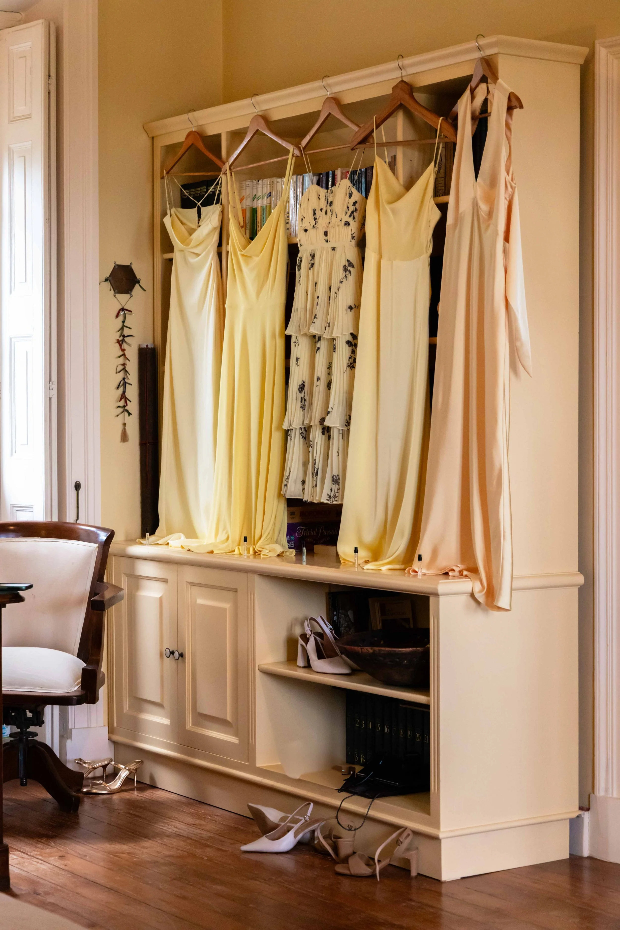 Formal yellow and floral dresses hanging from a white wooden wardrobe, with shoes on the floor and a cabinet with shelves underneath.