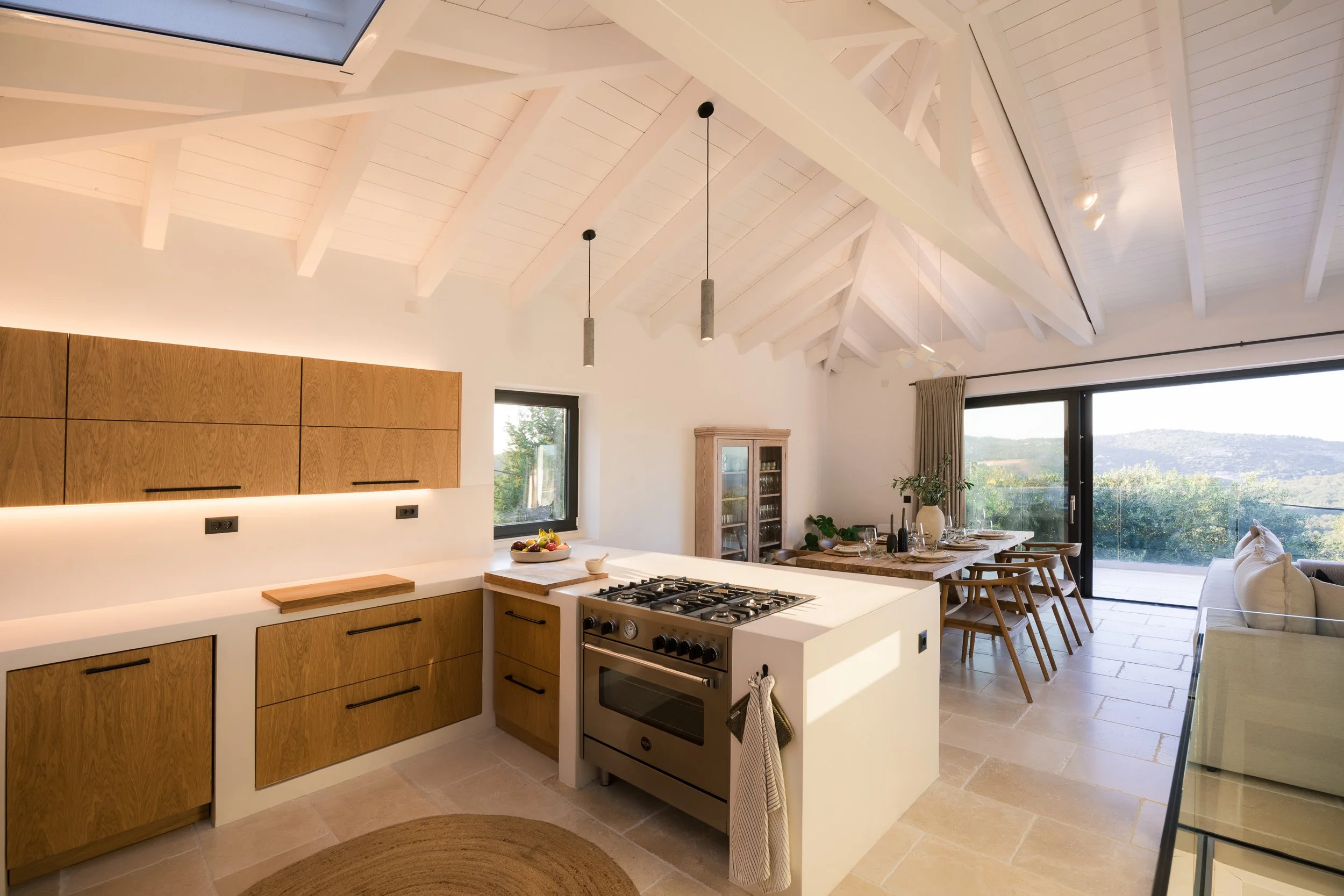 Open-concept kitchen and dining area with white vaulted ceiling, wooden cabinets, and large glass sliding door overlooking a scenic landscape.
