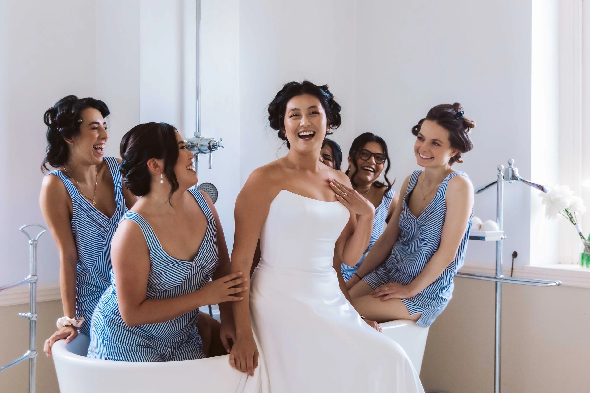 Bride and bridesmaids sitting on a white chair, laughing and smiling, in a bright room with white walls and large window, getting ready for a wedding.