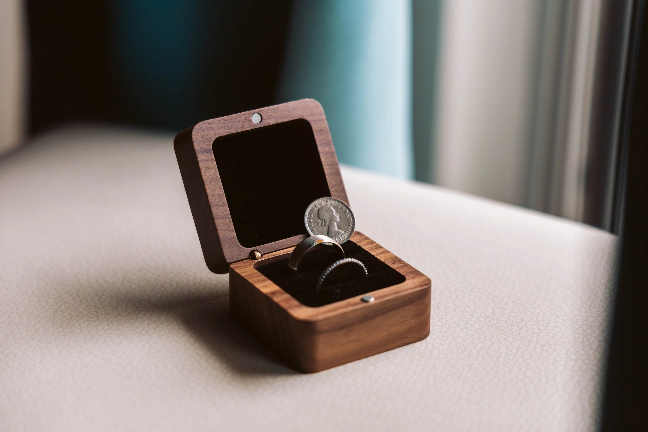 Wooden box containing a ring and a quarter coin on a light-colored surface near a window.