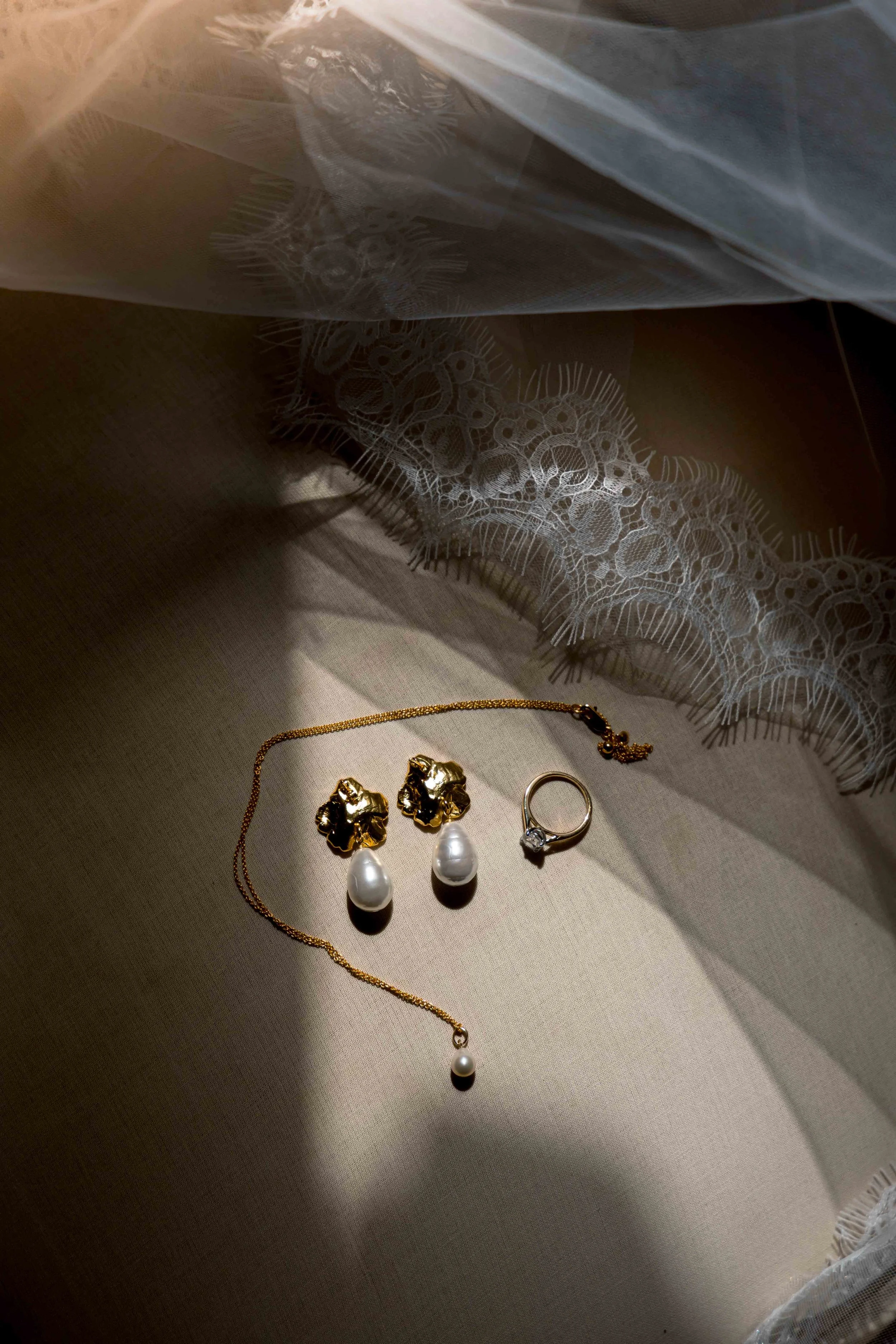 Jewelry including a gold necklace with a pearl pendant, a pair of gold earrings with pearl drops, and a ring with a small gemstone, arranged on a beige fabric surface with lace fabric in the background.
