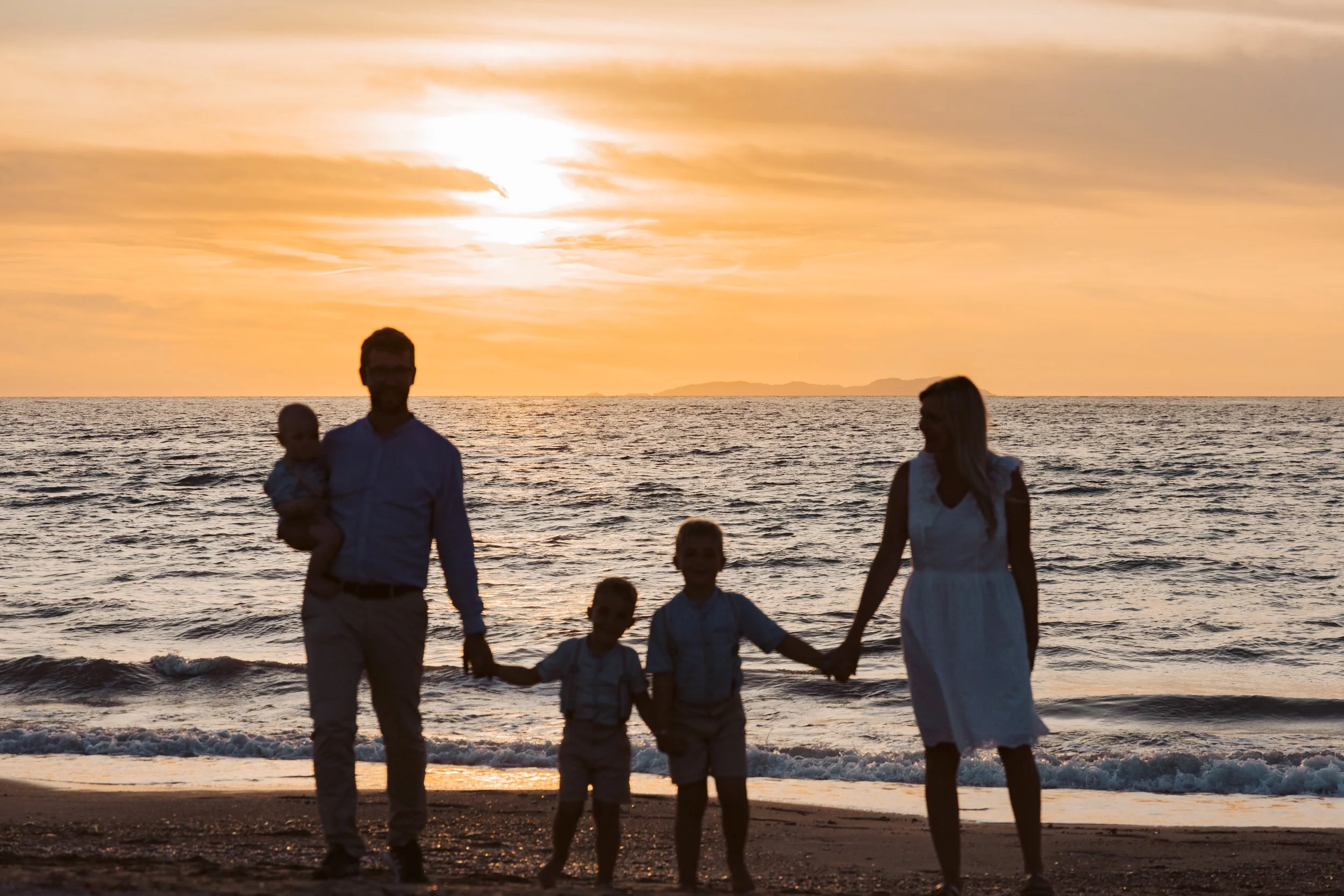 Silhouetted family of four holding hands on a beach at sunset, with a man holding a baby and a woman holding hands with two children, against a colorful sky and ocean background.