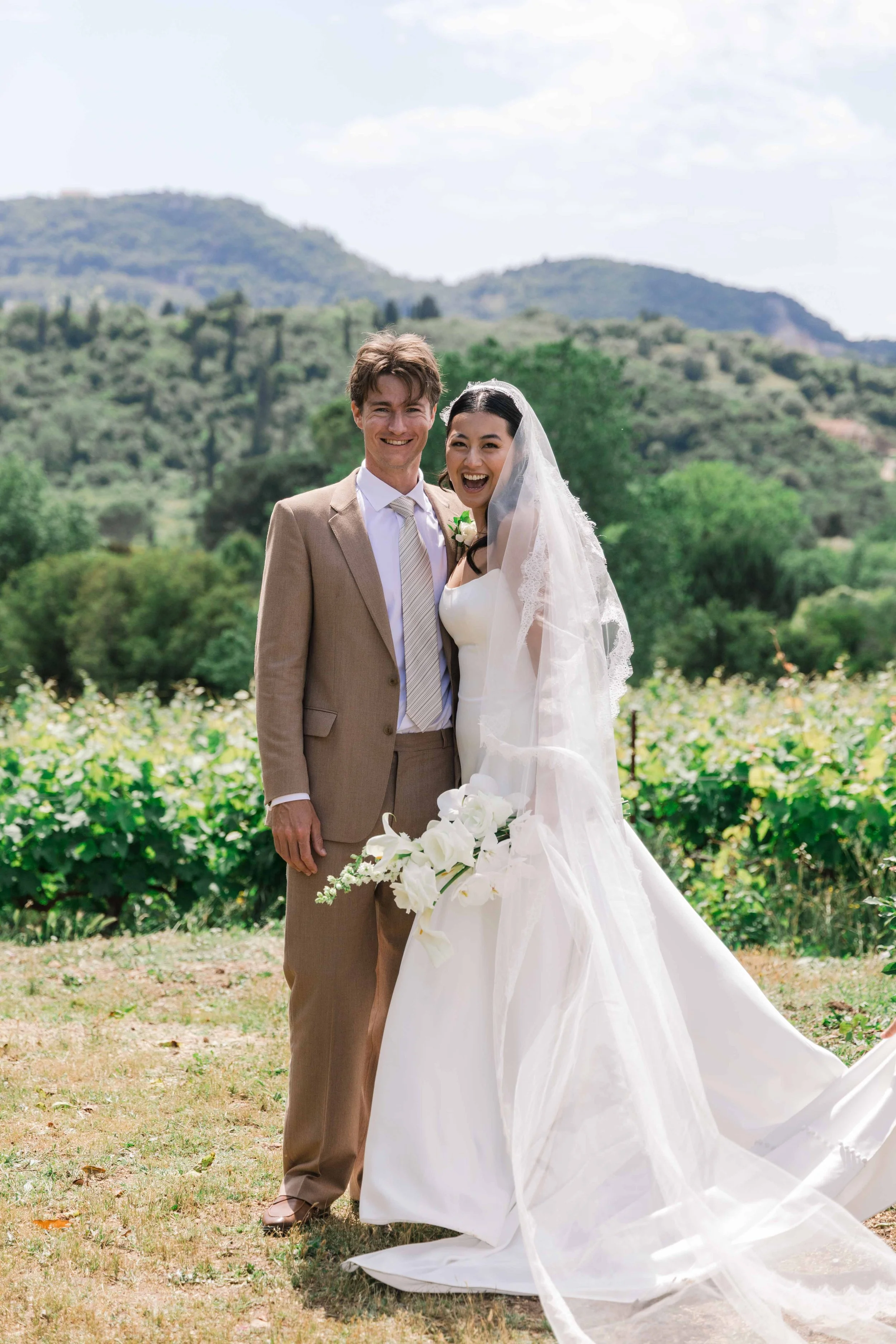 A newlywed couple standing outdoors on their wedding day, smiling. The bride is wearing a white wedding dress with a veil and holding a bouquet of white flowers. The groom is dressed in a beige suit with a tie. They are in a scenic setting with green