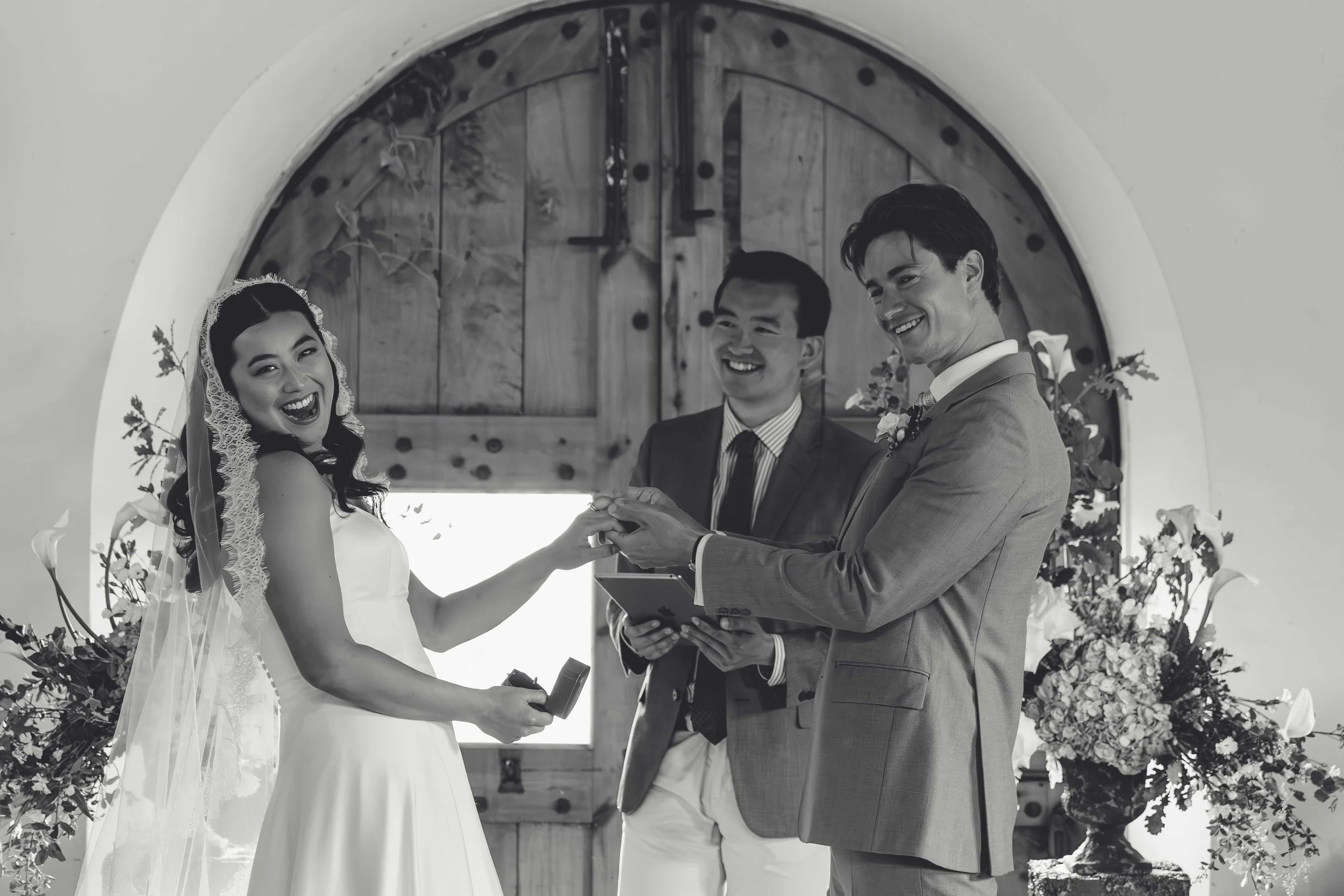 A wedding ceremony with a bride and groom exchanging rings, officiant presiding, in front of a large arched wooden door with floral arrangements.