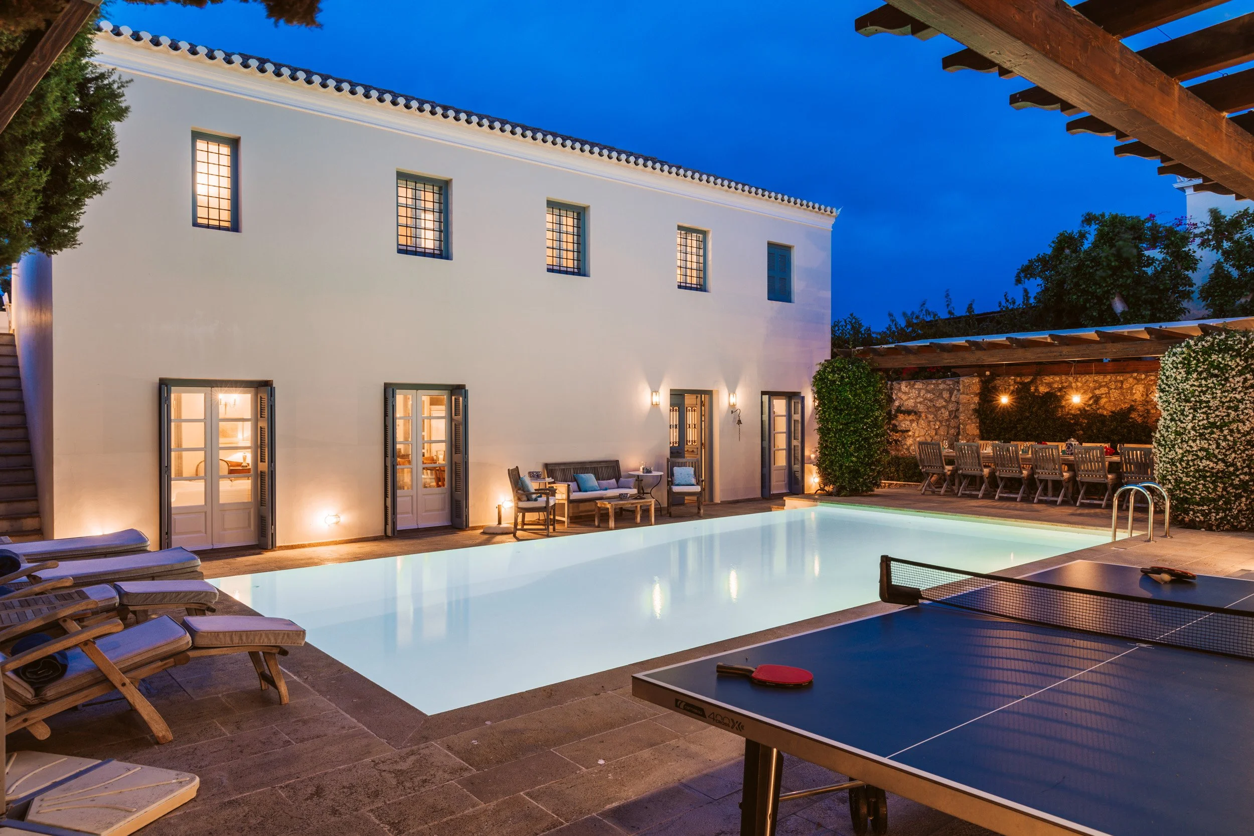 A backyard with a lit rectangular swimming pool, surrounded by wooden lounge chairs, a ping pong table, outdoor seating, and string lights on a stone patio at dusk, with a white house and greenery in the background.