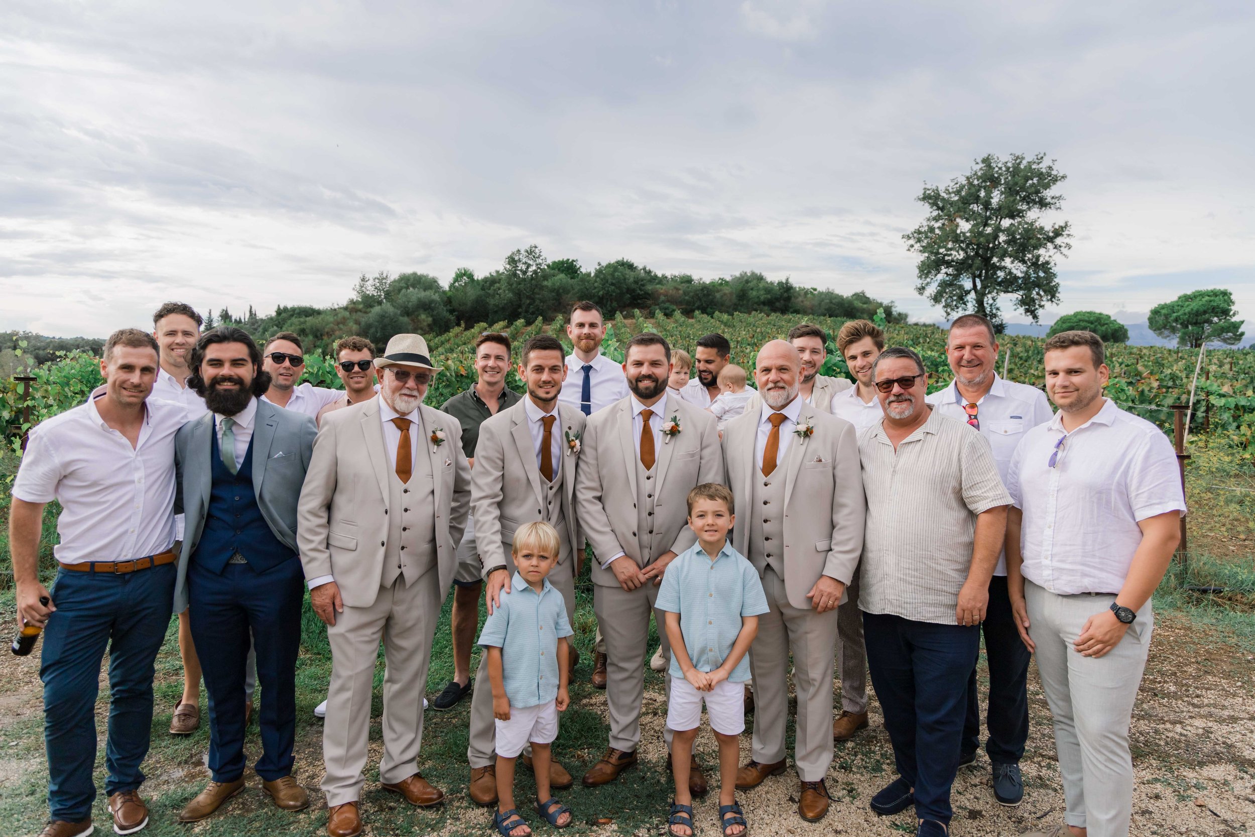 Group of men, women, and children standing outdoors in a vineyard with hills and trees in the background, dressed for a wedding.