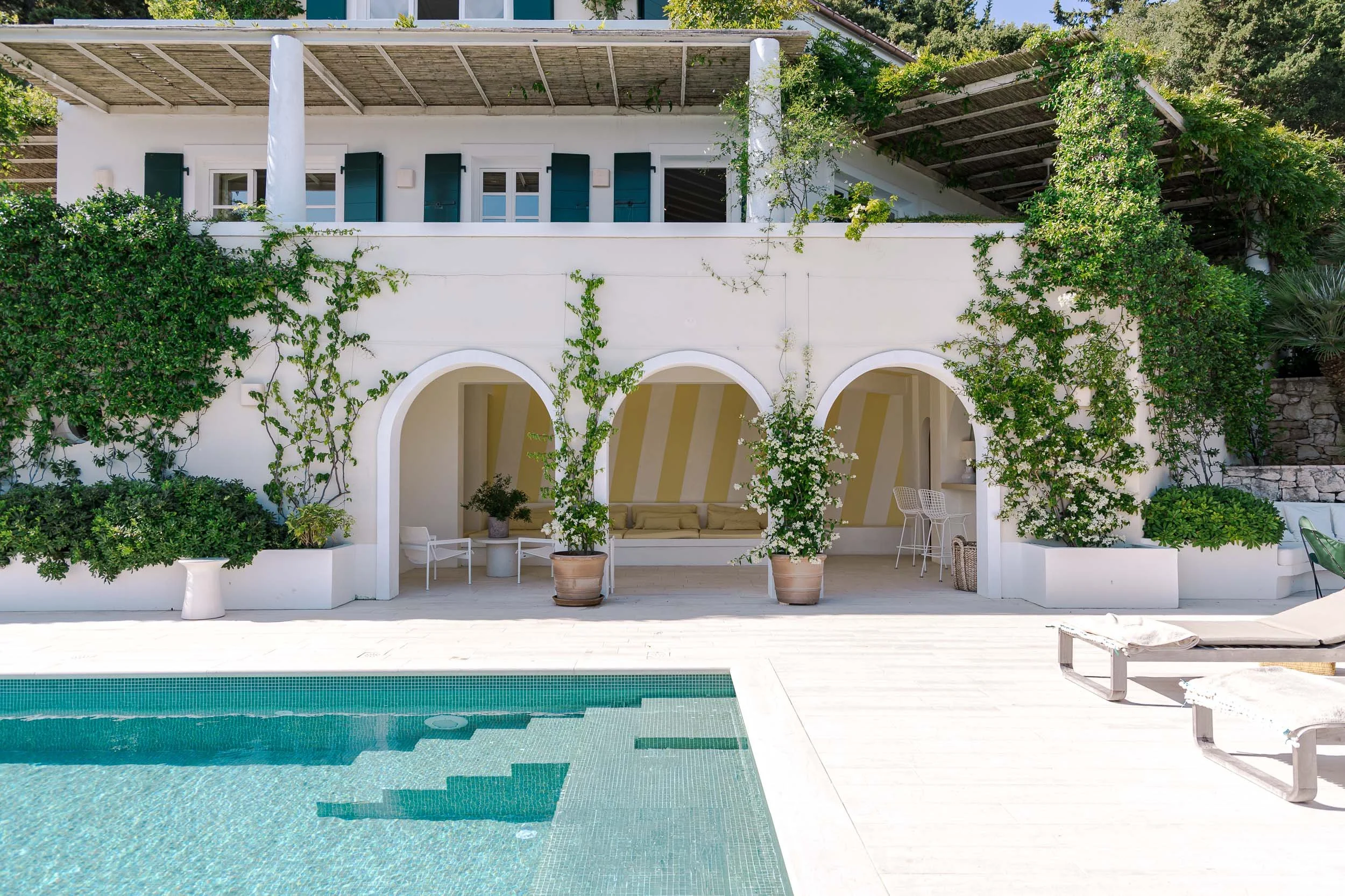 A white house with arched doorways and green shutters, greenery growing on the walls, a pool in the foreground, and a striped covered patio area with outdoor furniture.