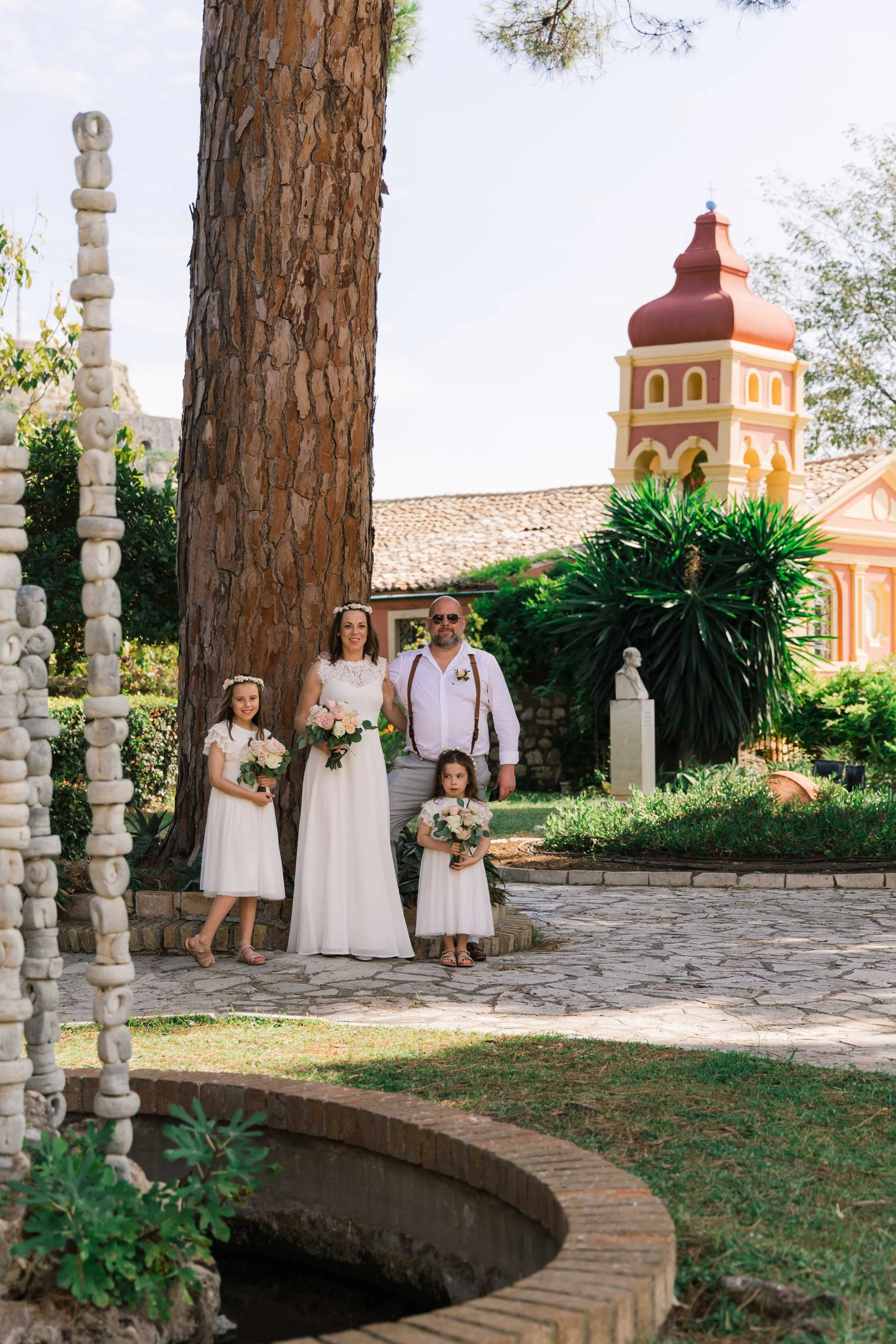 A family of five standing outdoors in a garden, dressed in wedding attire. The woman and three young girls are holding bouquets, and the man is wearing sunglasses with suspenders. They are standing next to a large tree with a church or chapel buildin
