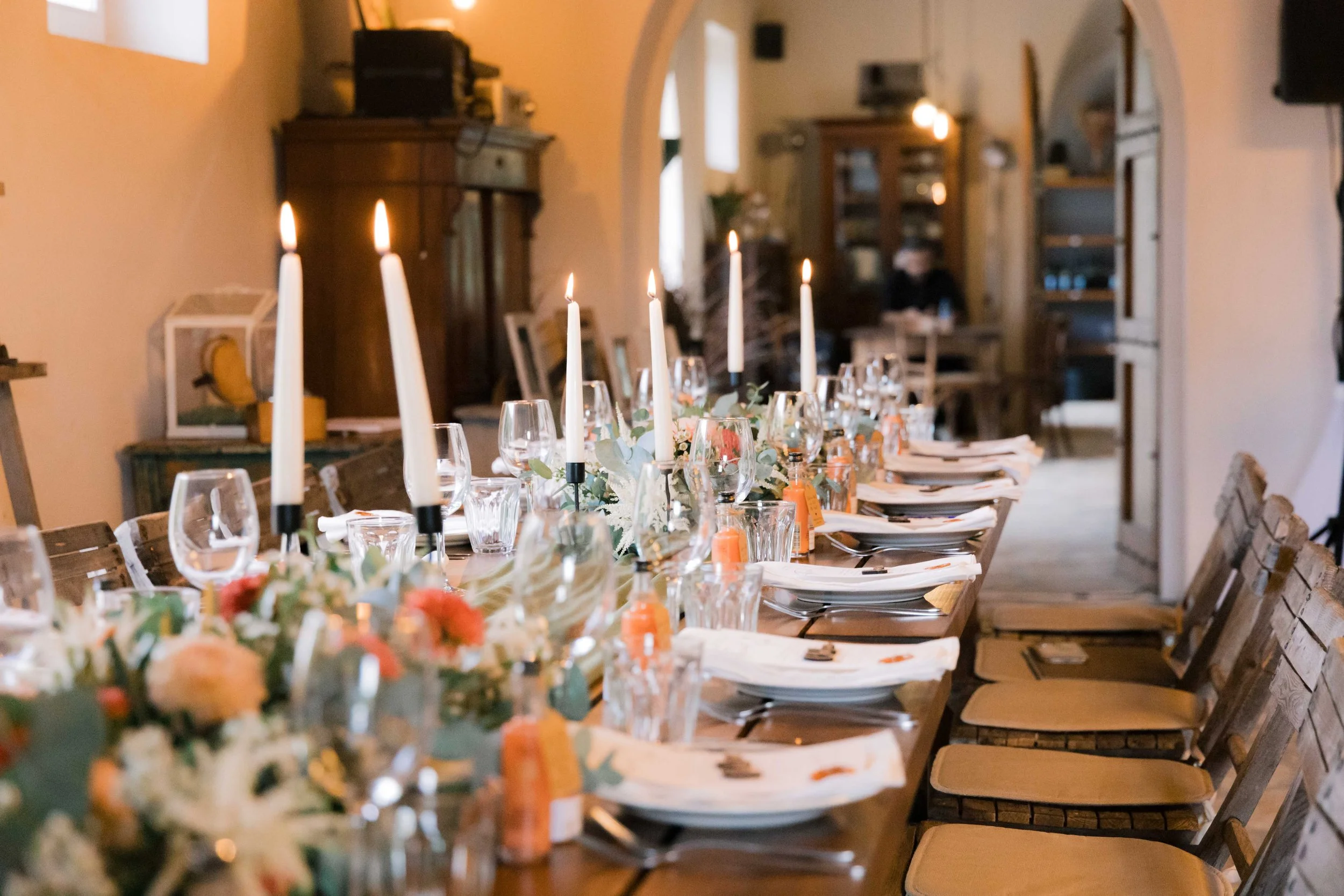 A long dining table set for a formal dinner with white candles, floral arrangements, wine glasses, plates, napkins, and chairs in a warmly lit rustic dining room.