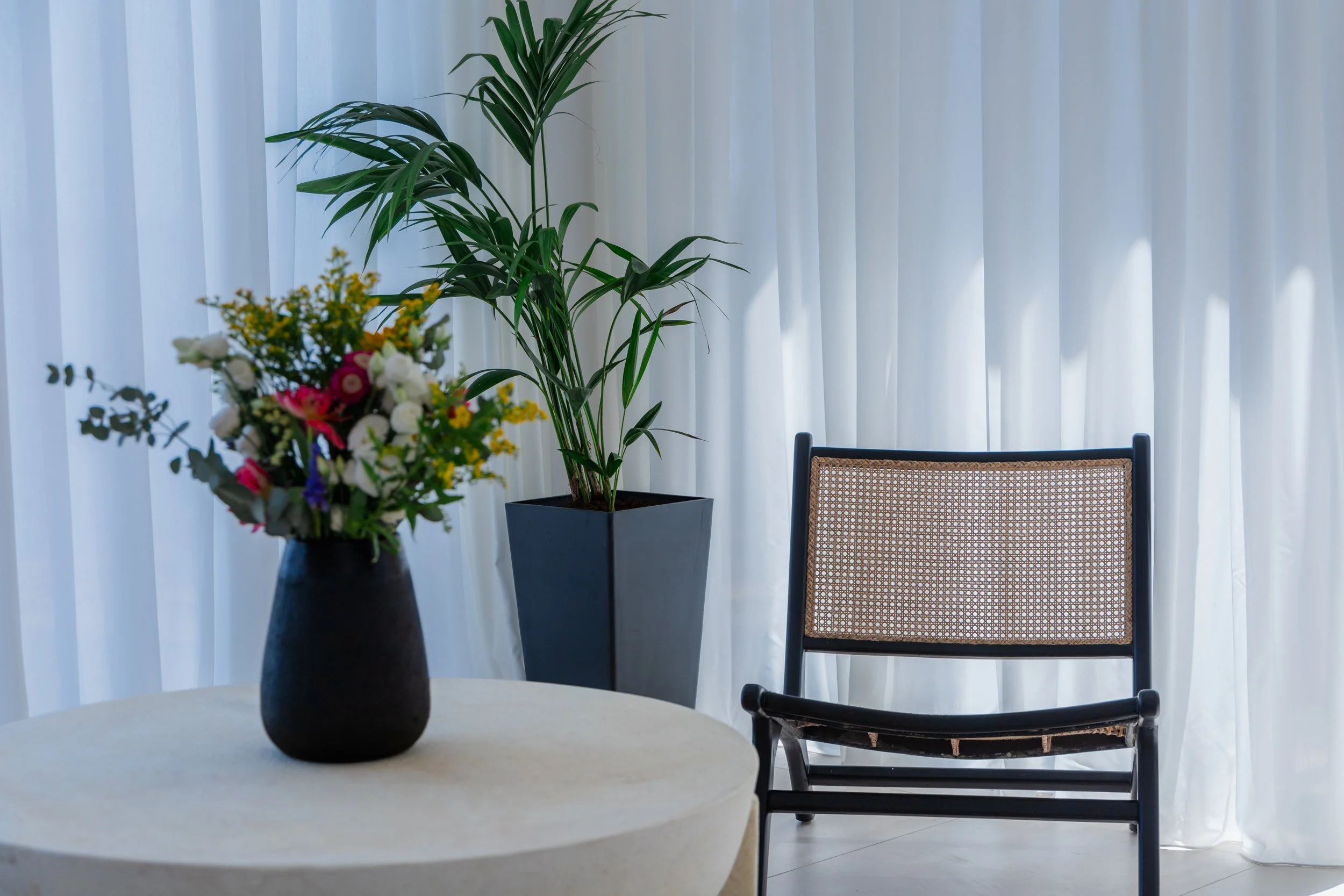 A living room with a white curtain, a black chair with a rattan seat and backrest, a large potted plant with green leaves, and a bouquet of colorful flowers on a round table.