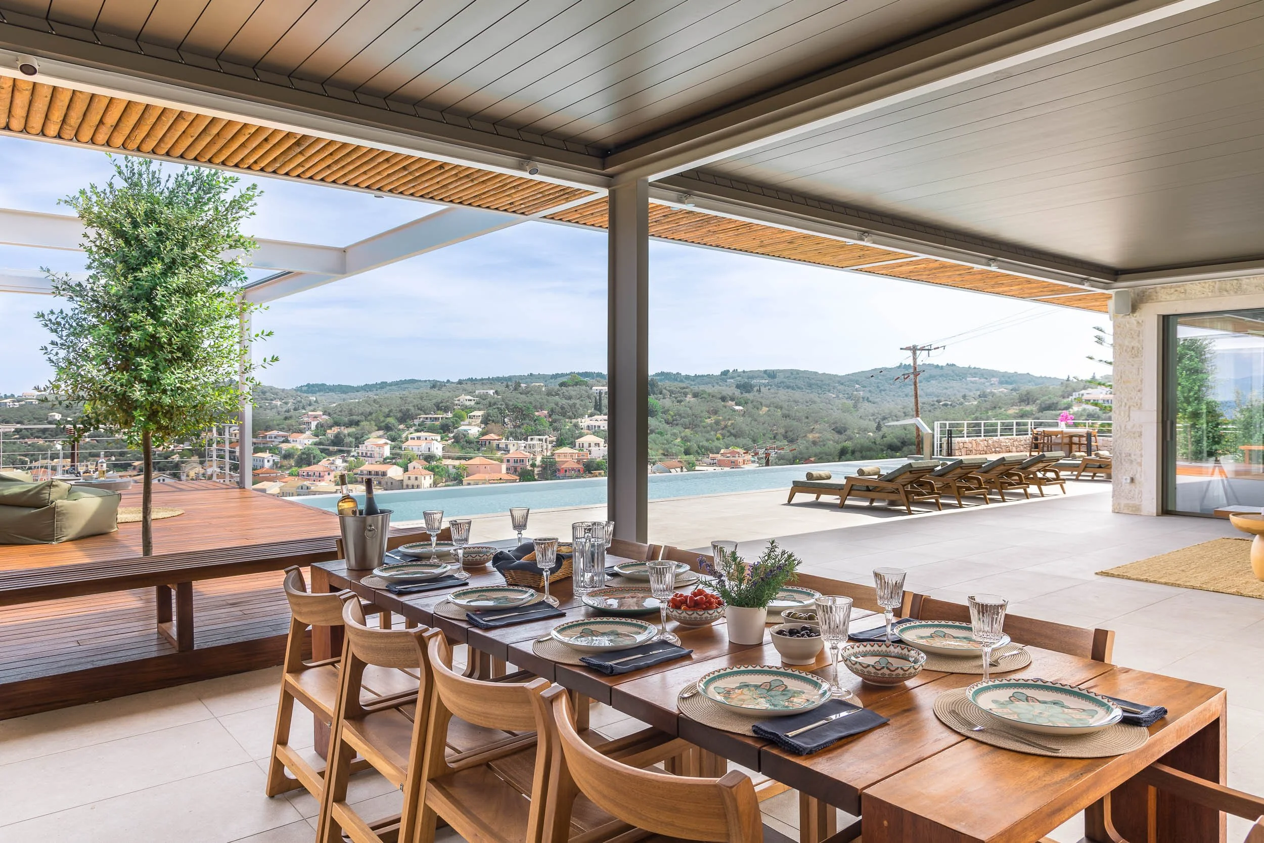 Outdoor terrace with a wooden dining table set with plates, glasses, and a potted plant, overlooking a pool, lounge chairs, and a scenic hillside view.