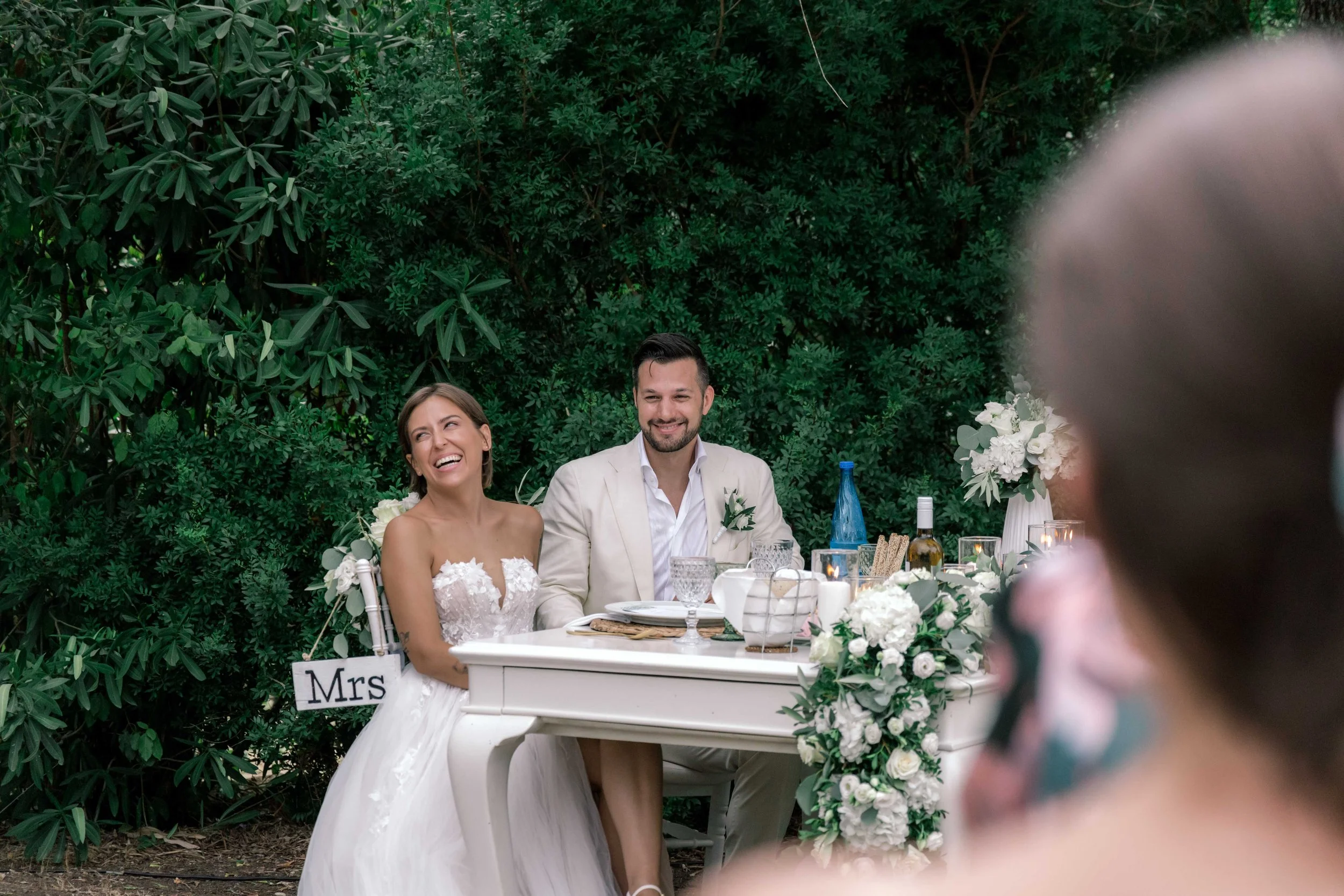 A wedding reception outdoors with a bride and groom seated at a white table decorated with white flowers and greenery. The bride is wearing a white wedding dress and laughing, while the groom is in a light-colored suit, smiling. A guest in the foregr