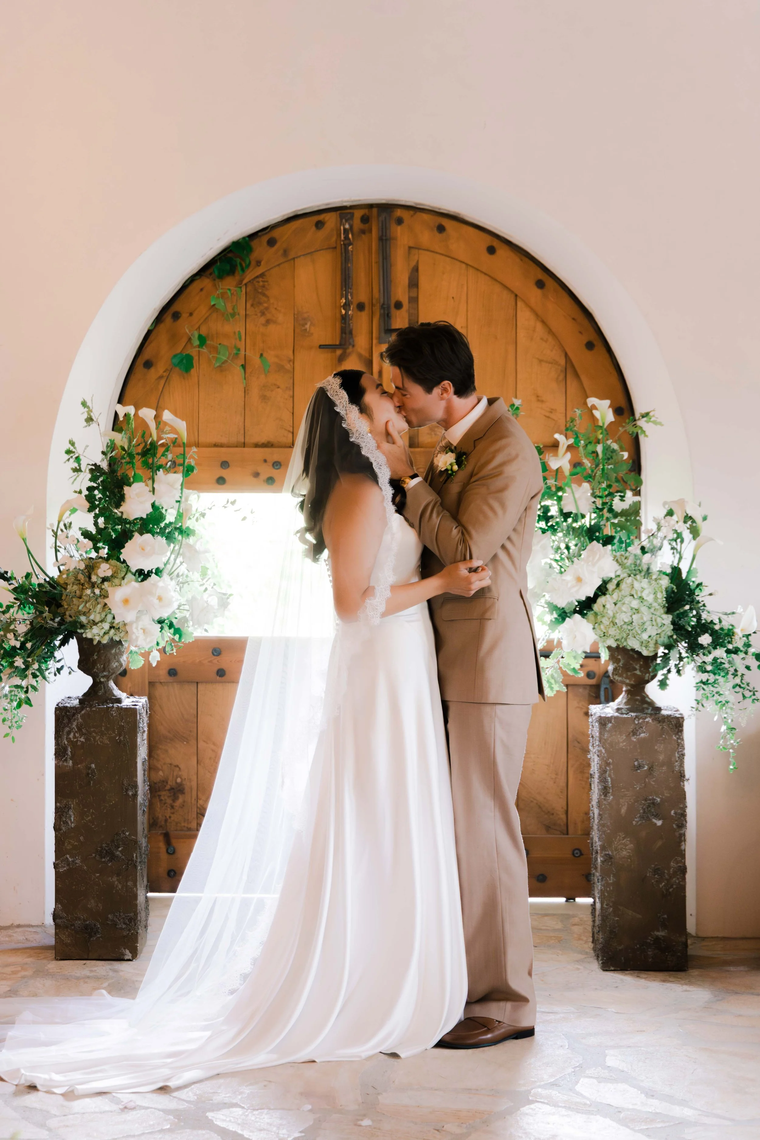 A bride and groom sharing a kiss in front of a wooden door with floral arrangements on each side.