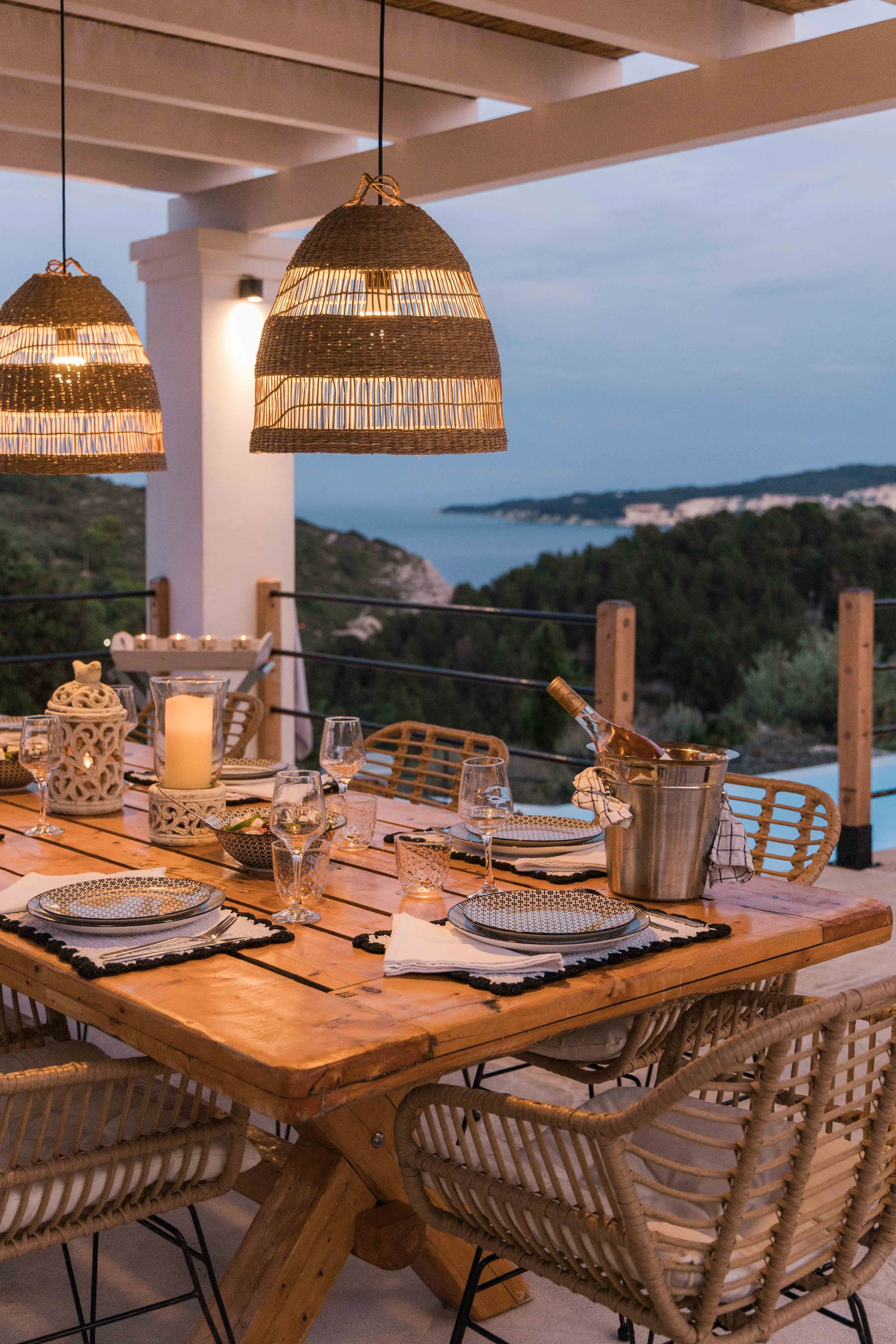 An outdoor dining table set for a meal at dusk, with wicker chairs, decorative lanterns, candles, wine glasses, and a bottle of wine in an ice bucket. The table is on a balcony with a view of trees, hills, and the ocean.