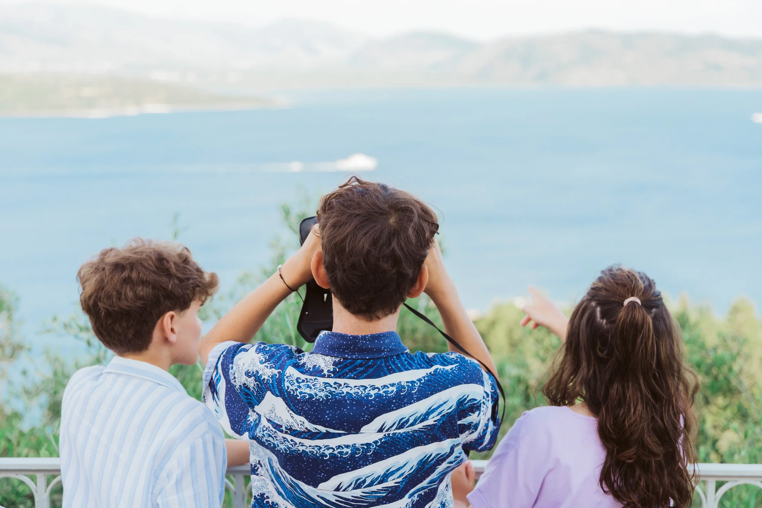 Three children looking out at a large blue body of water with boats, on a hillside with green trees in the foreground.