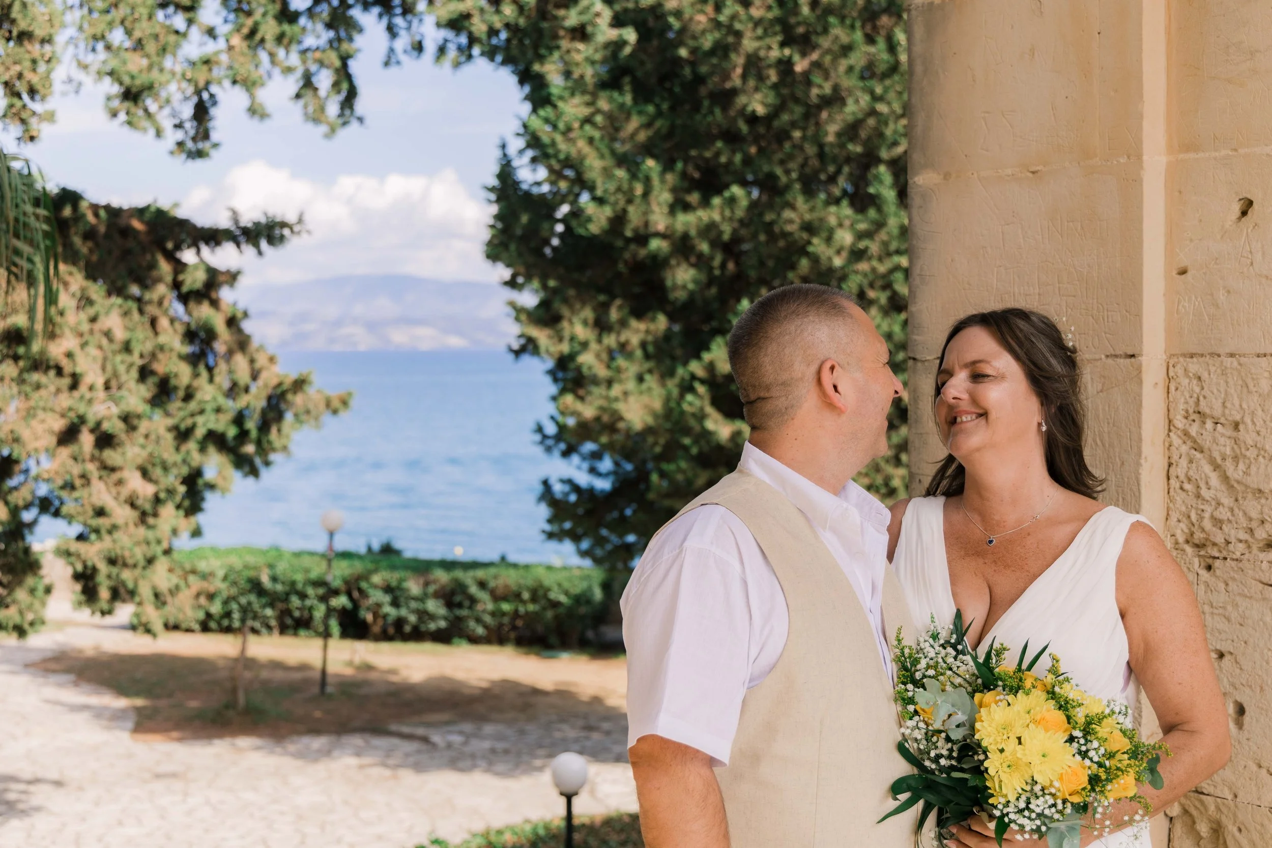 A couple dressed in wedding attire standing outdoors near a stone wall, smiling at each other with a lake and trees in the background, holding a bouquet of yellow and white flowers.