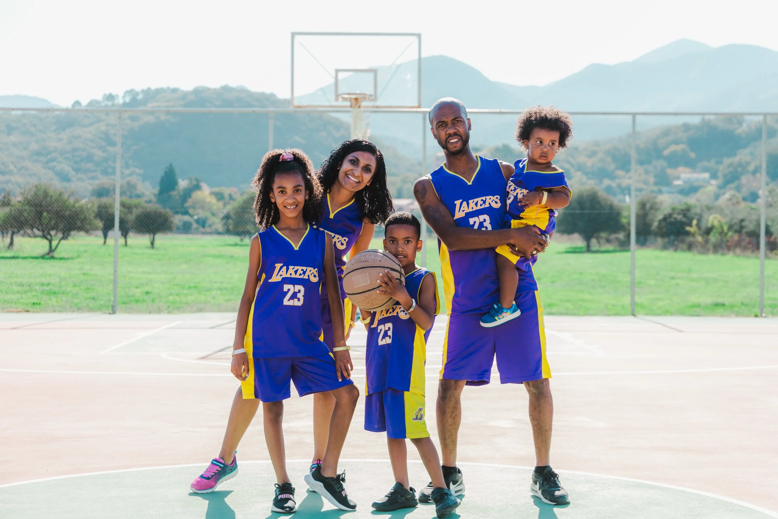 A family of five dressed in blue and yellow Lakers basketball jerseys, standing on an outdoor basketball court with mountains in the background.