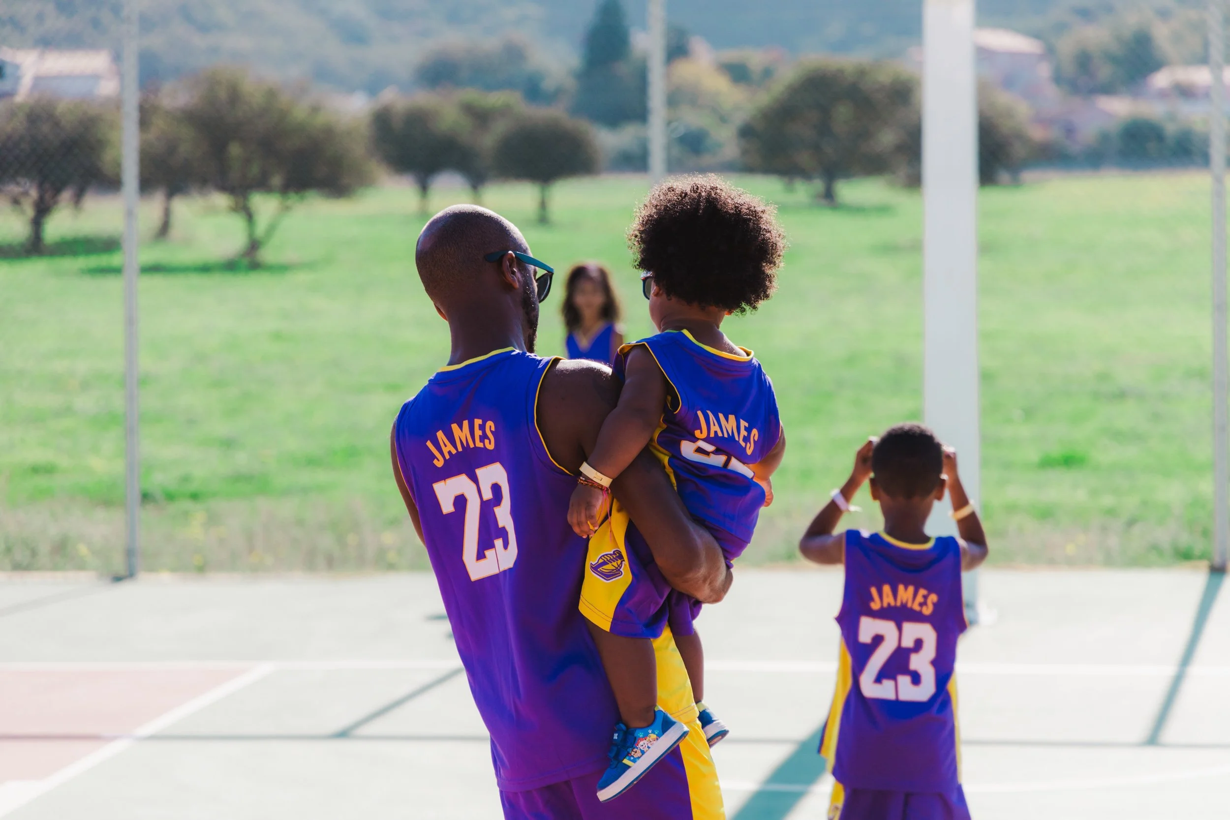 A man holding a child and a boy on a basketball court, all wearing purple and yellow jerseys with the name 'James' and the number 23.