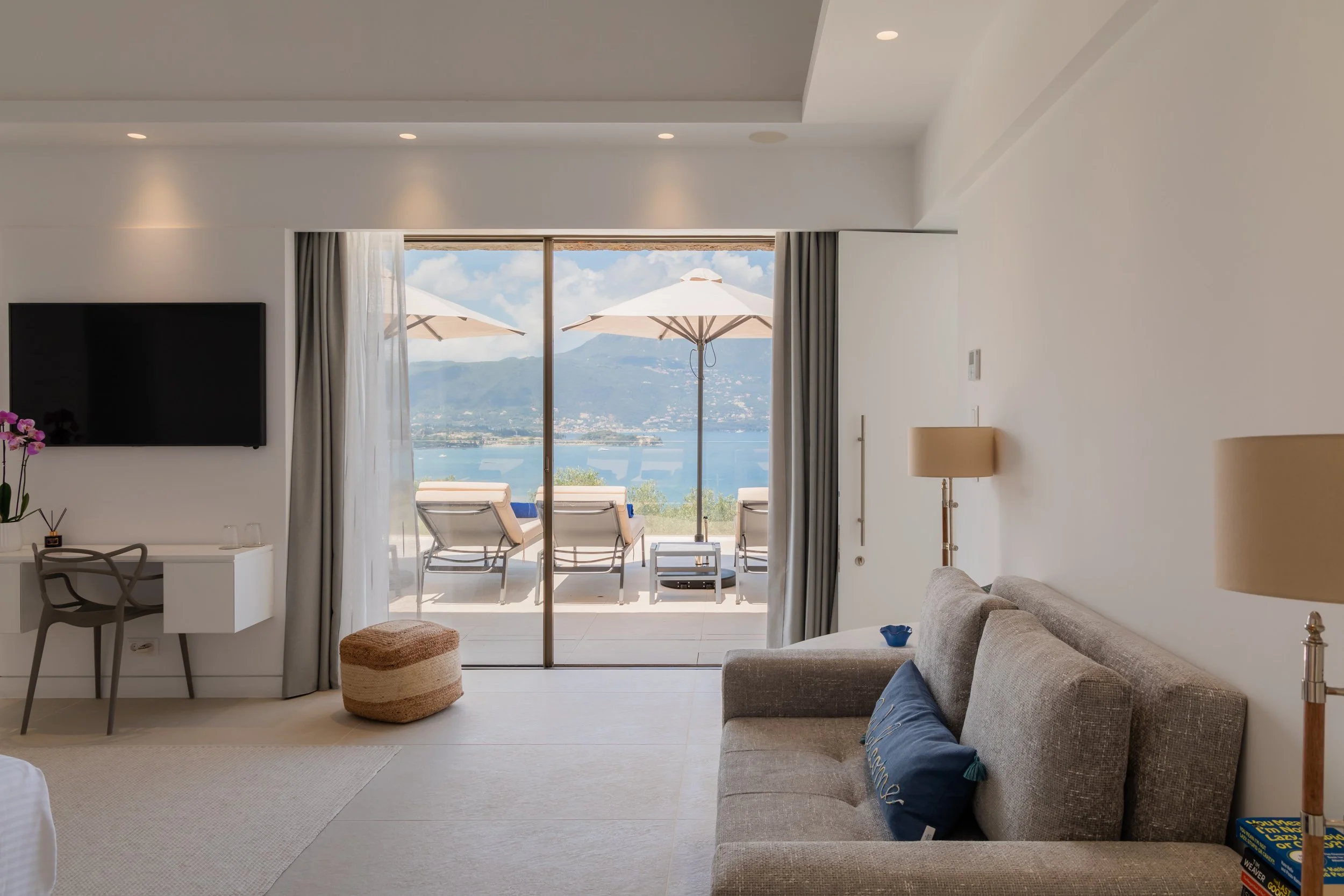 Living room with a view of an outdoor patio and water beyond, featuring lounge chairs, umbrellas, a flat-screen TV, a desk with a chair, a sofa with pillows, side lamps, and a sliding glass door opening to the patio.