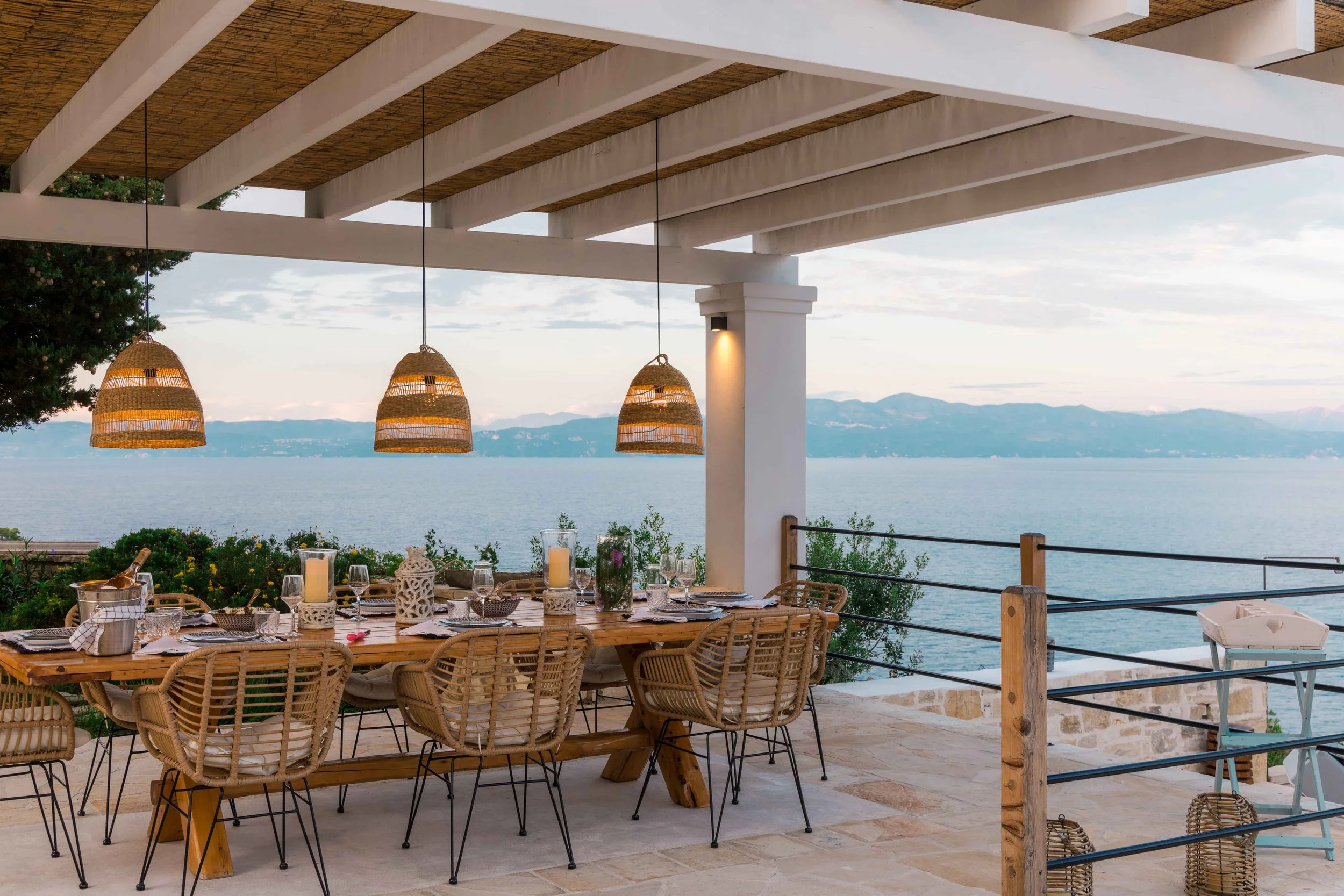 Outdoor dining area on a terrace overlooking a large body of water with mountains in the distance. The table is set with candles, candles, glasses, and tableware, and surrounded by rattan chairs. Woven pendant lights hang above the table.