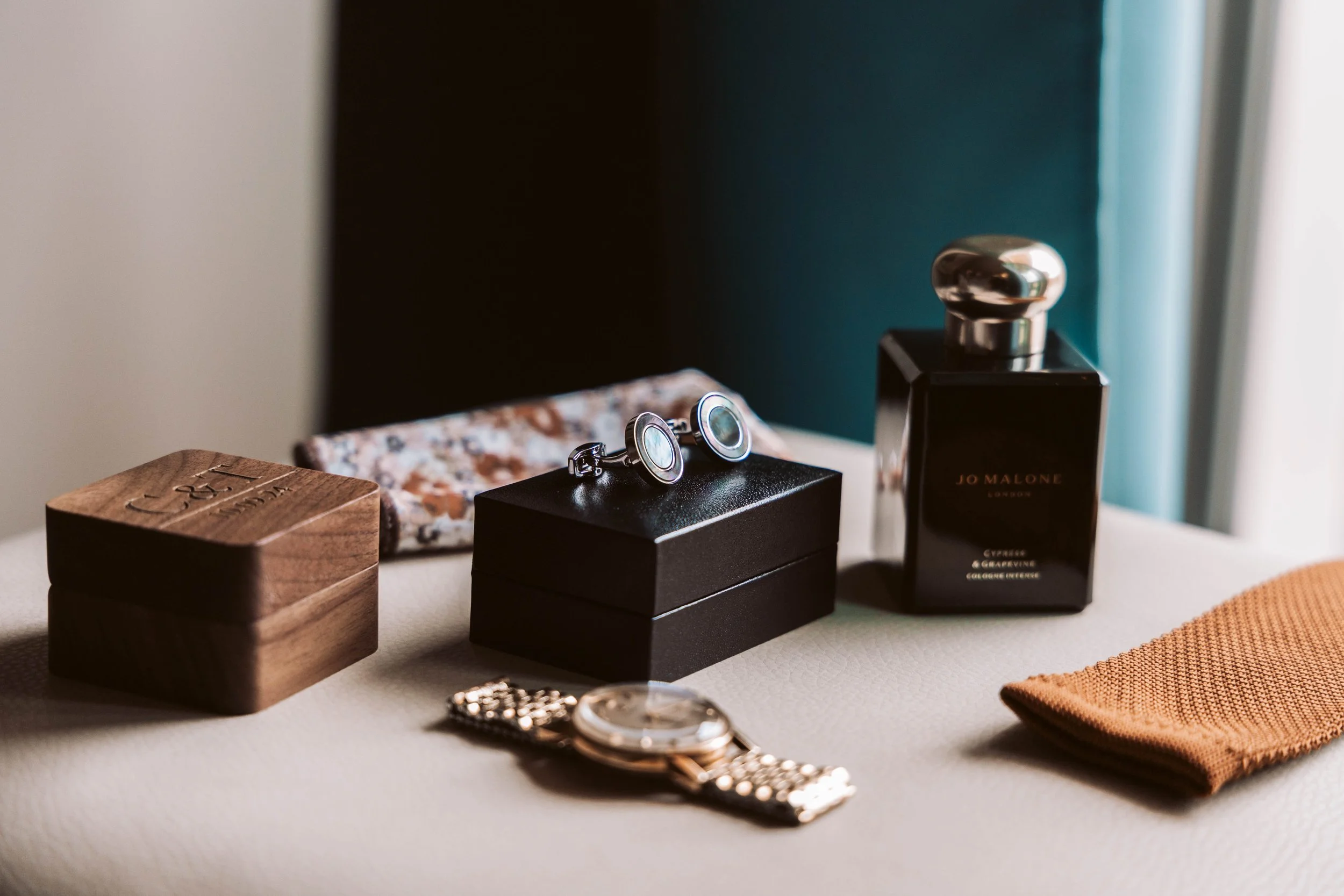 A collection of men's accessories on a white surface, including a wooden box, a floral-patterned glasses case, a pair of cufflinks on a black box, a watch with a metal strap, a bottle of Jo Malone perfume, and a brown textured cloth.