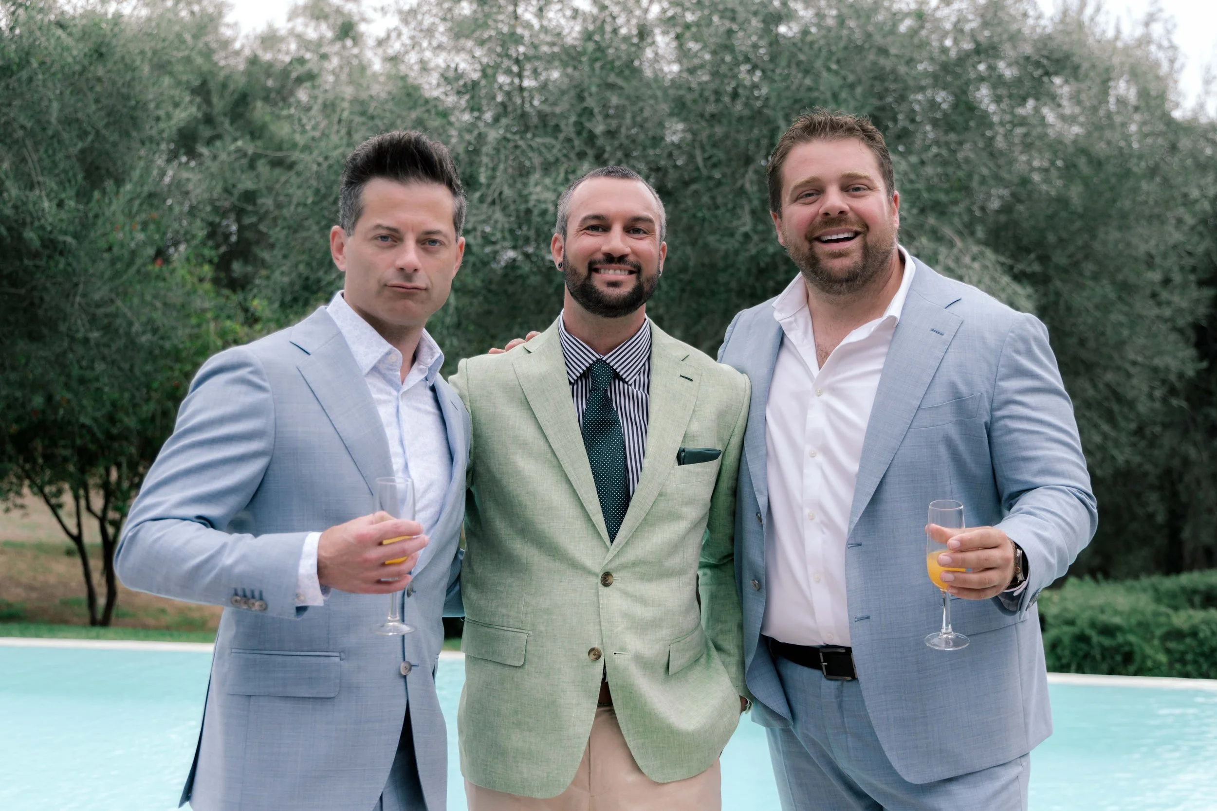 Three men in suits standing by a swimming pool, holding drinks, smiling at the camera, with greenery in the background.