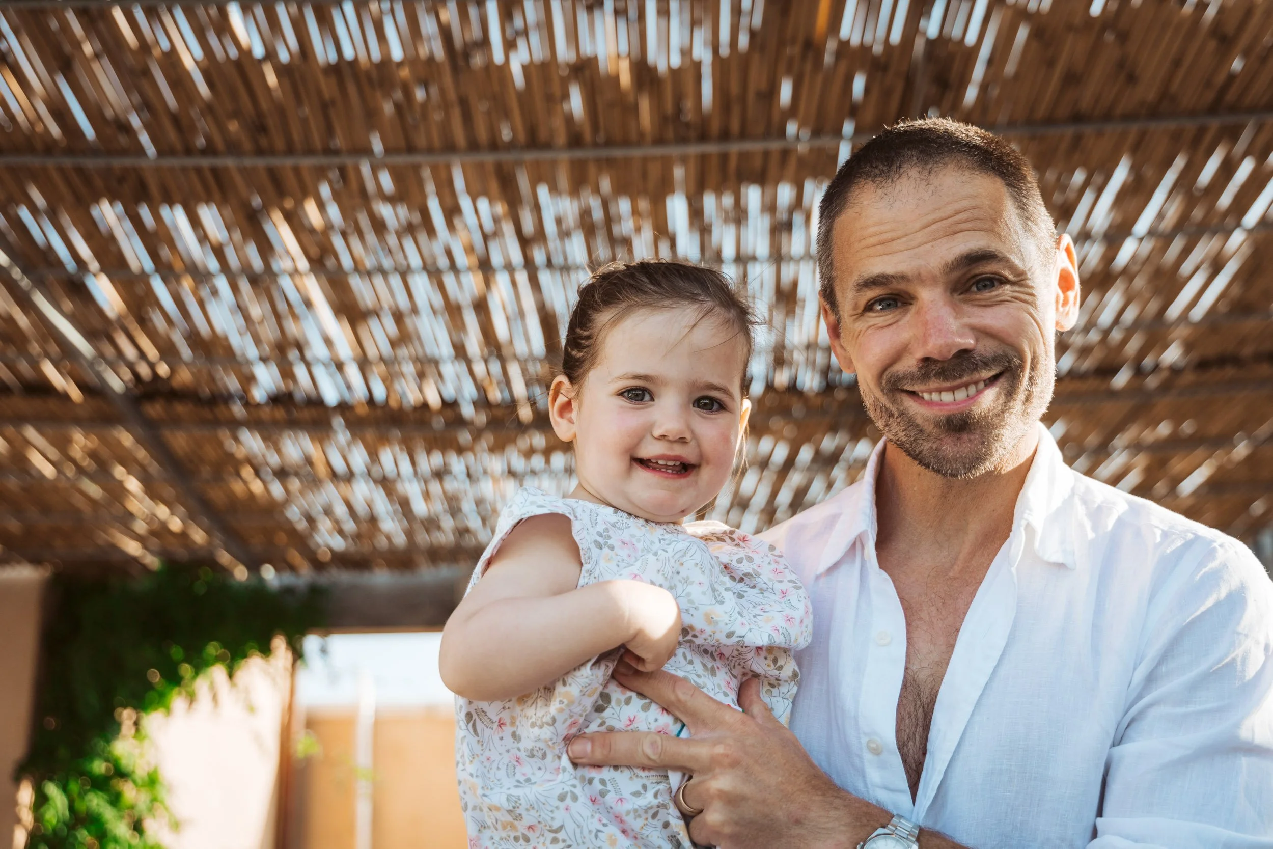 A smiling man holding a young girl under a wooden pergola with sunlight.