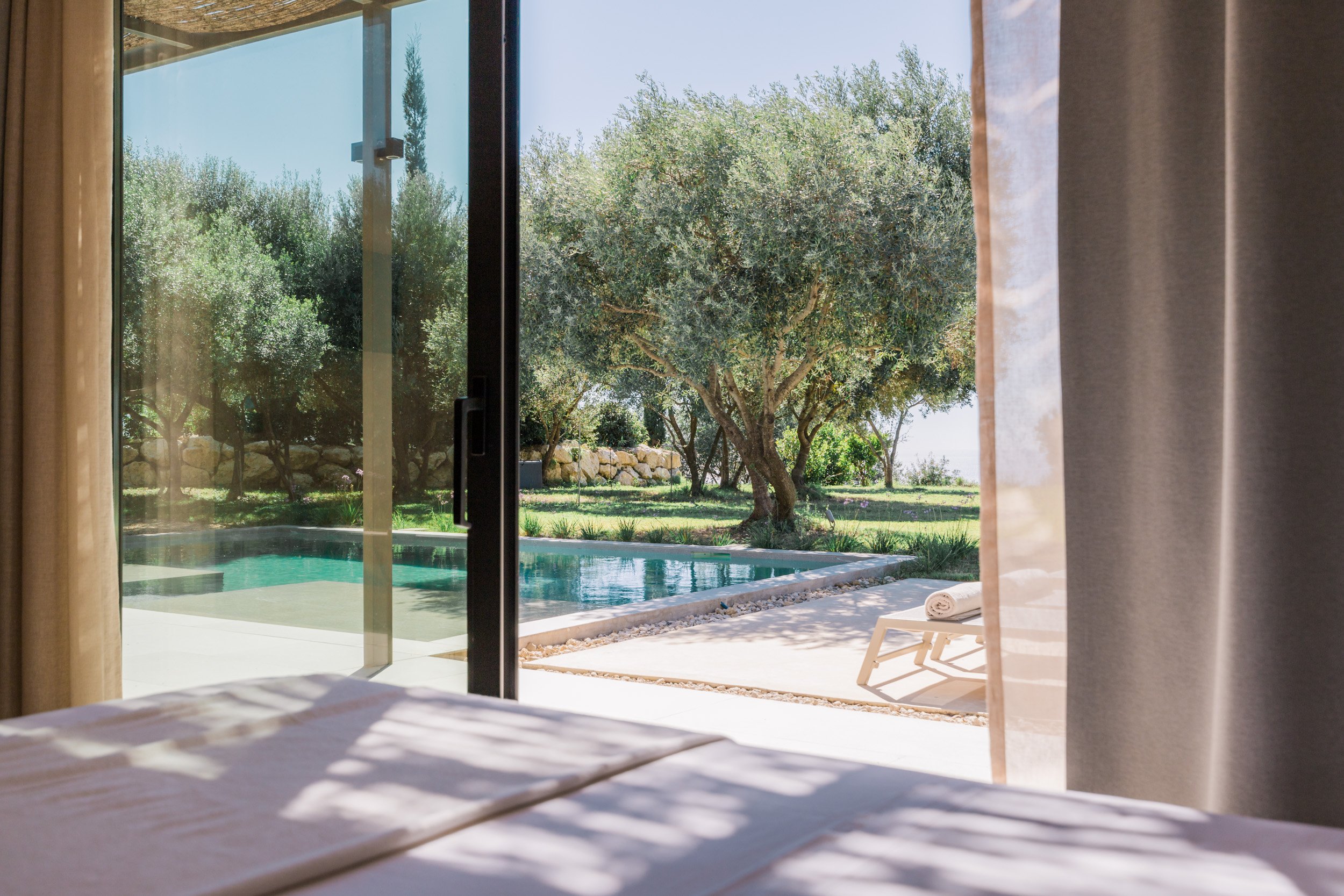 View through sliding glass door of backyard with swimming pool, trees, and lawn, with a white lounge chair and rolled towel outside.