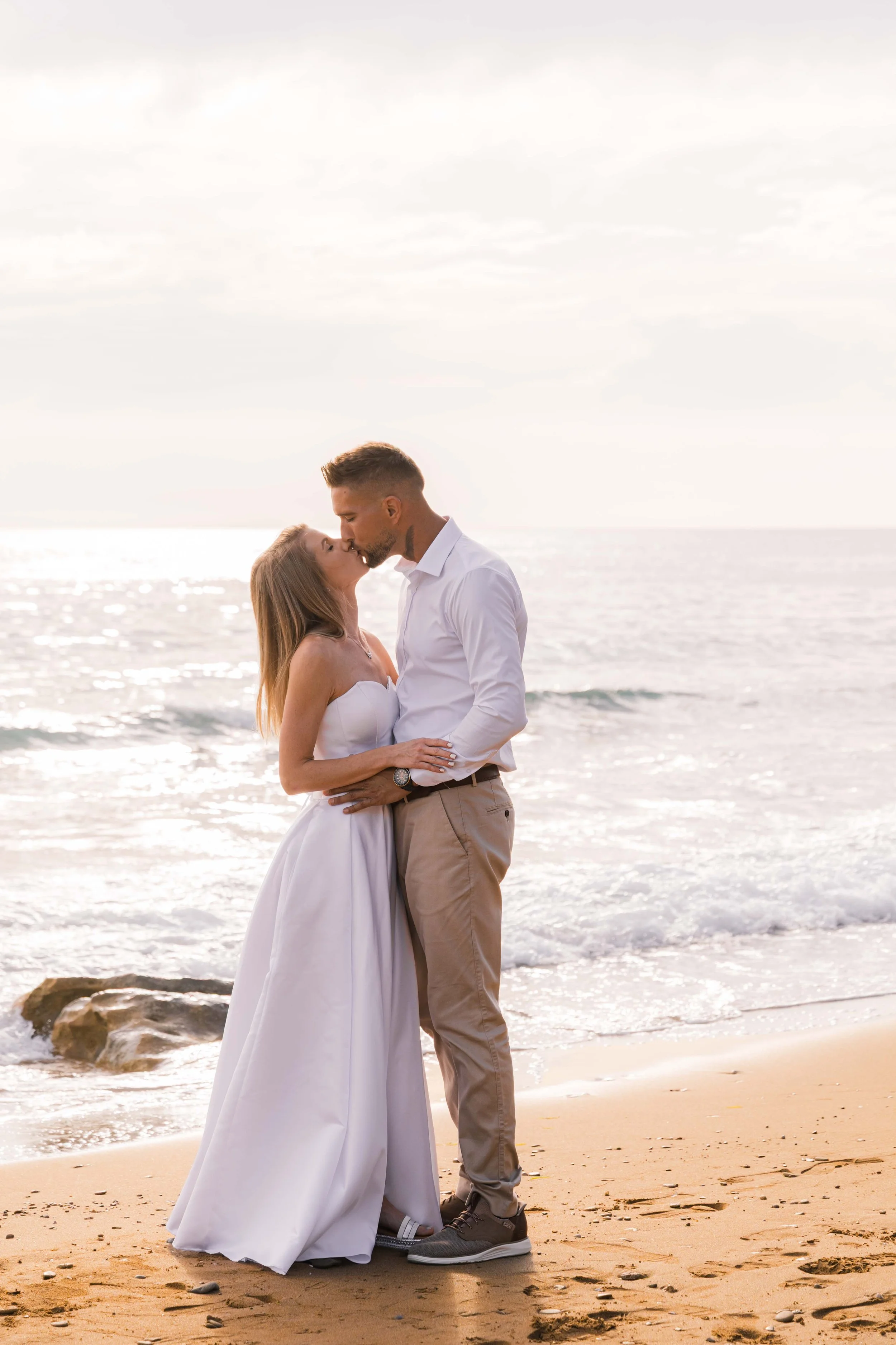 A couple sharing a romantic kiss on the beach at sunset, with the ocean in the background and footprints in the sand.