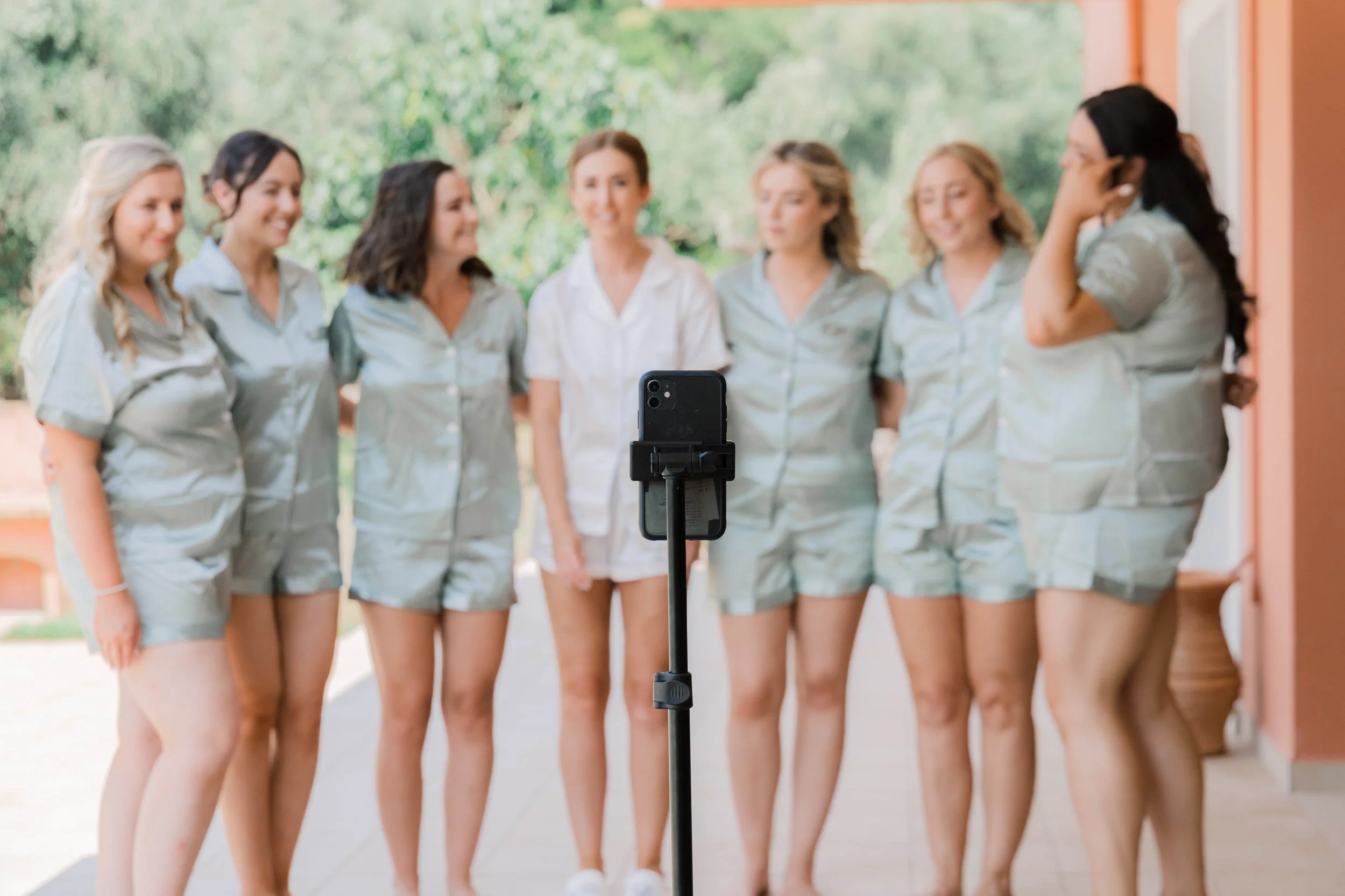 A group of seven women dressed in matching light-colored pajamas taking a group photo outdoors using a smartphone mounted on a selfie stick, with trees and a building in the background.
