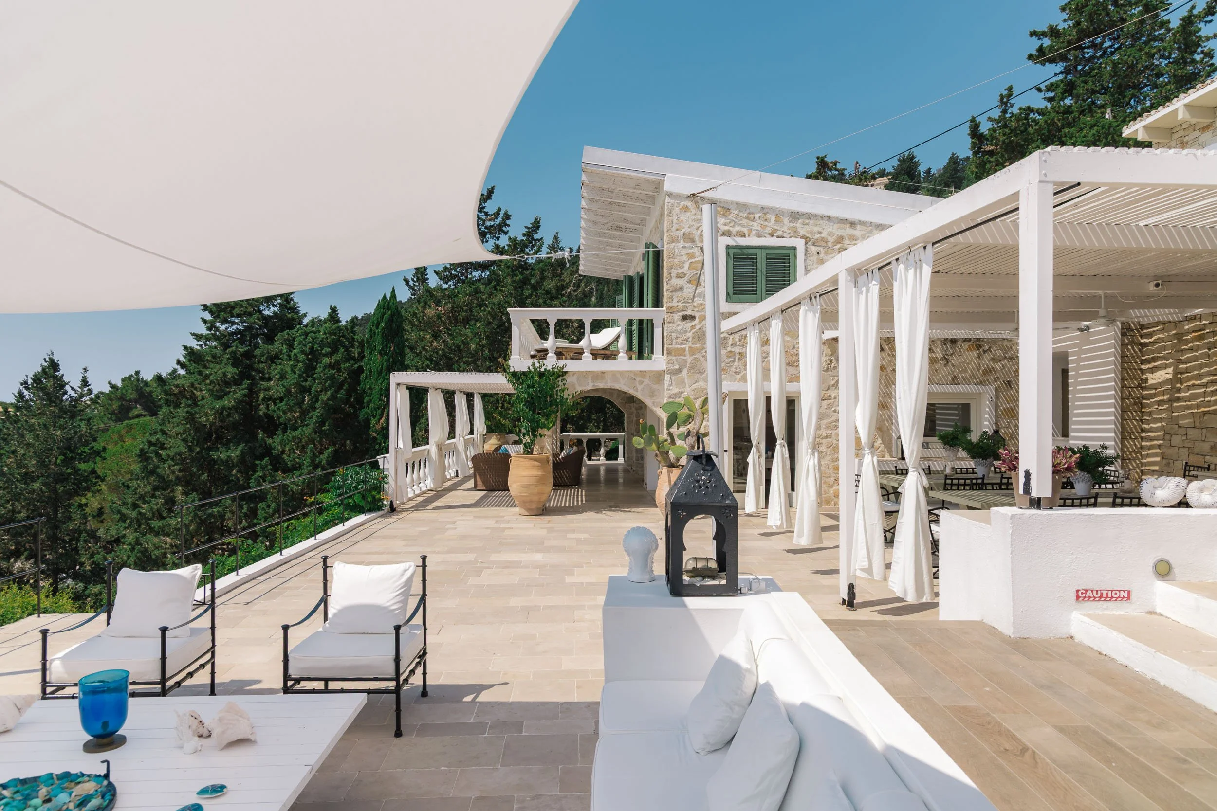 A spacious outdoor terrace of a Mediterranean-style house with stone walls and white accents. The terrace is partially covered with white curtains and has outdoor furniture including chairs, a table, and decorative items. There is greenery surroundin